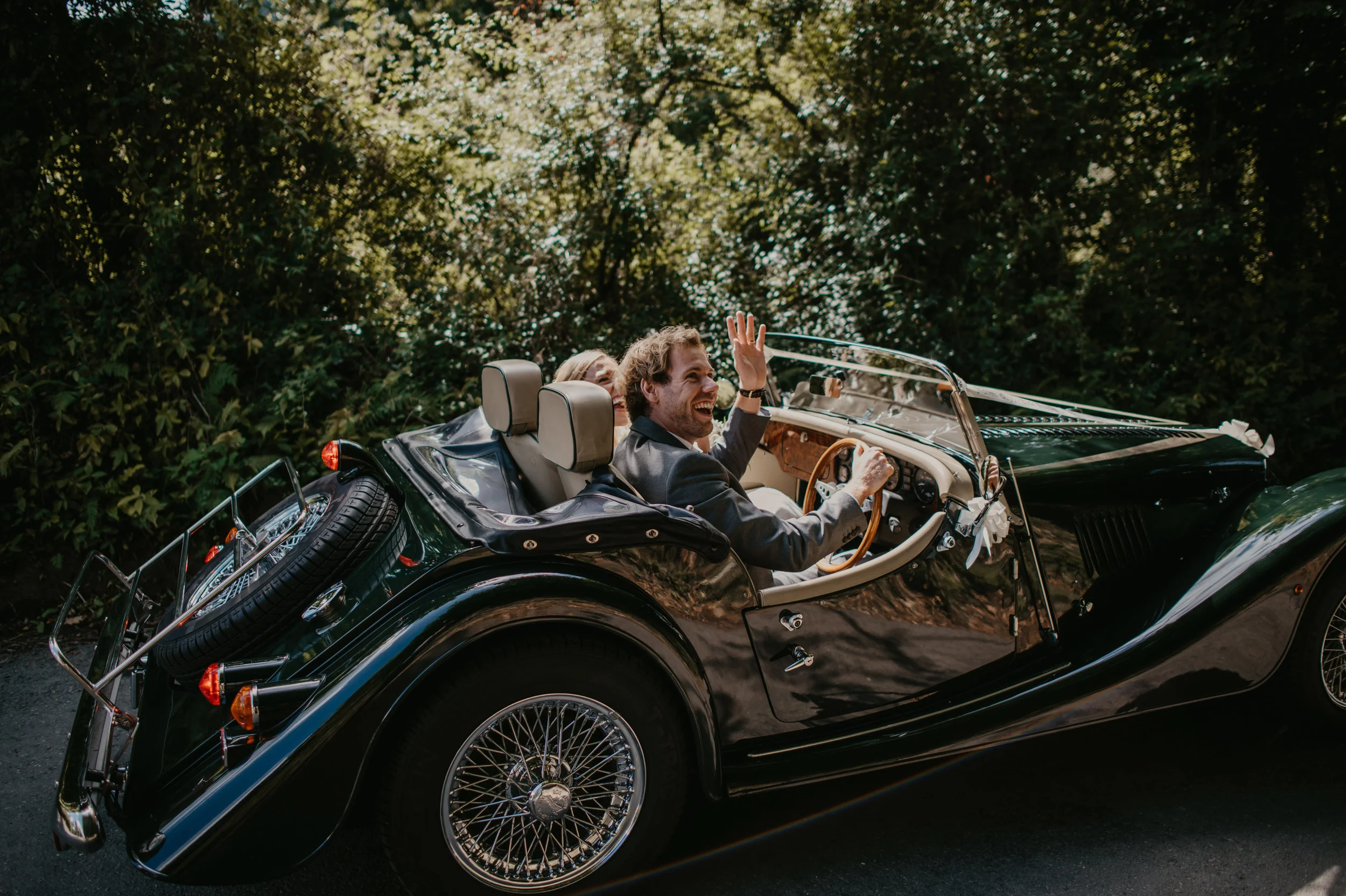 A man and woman smiling and waving from a classic black convertible car on a tree-lined road.