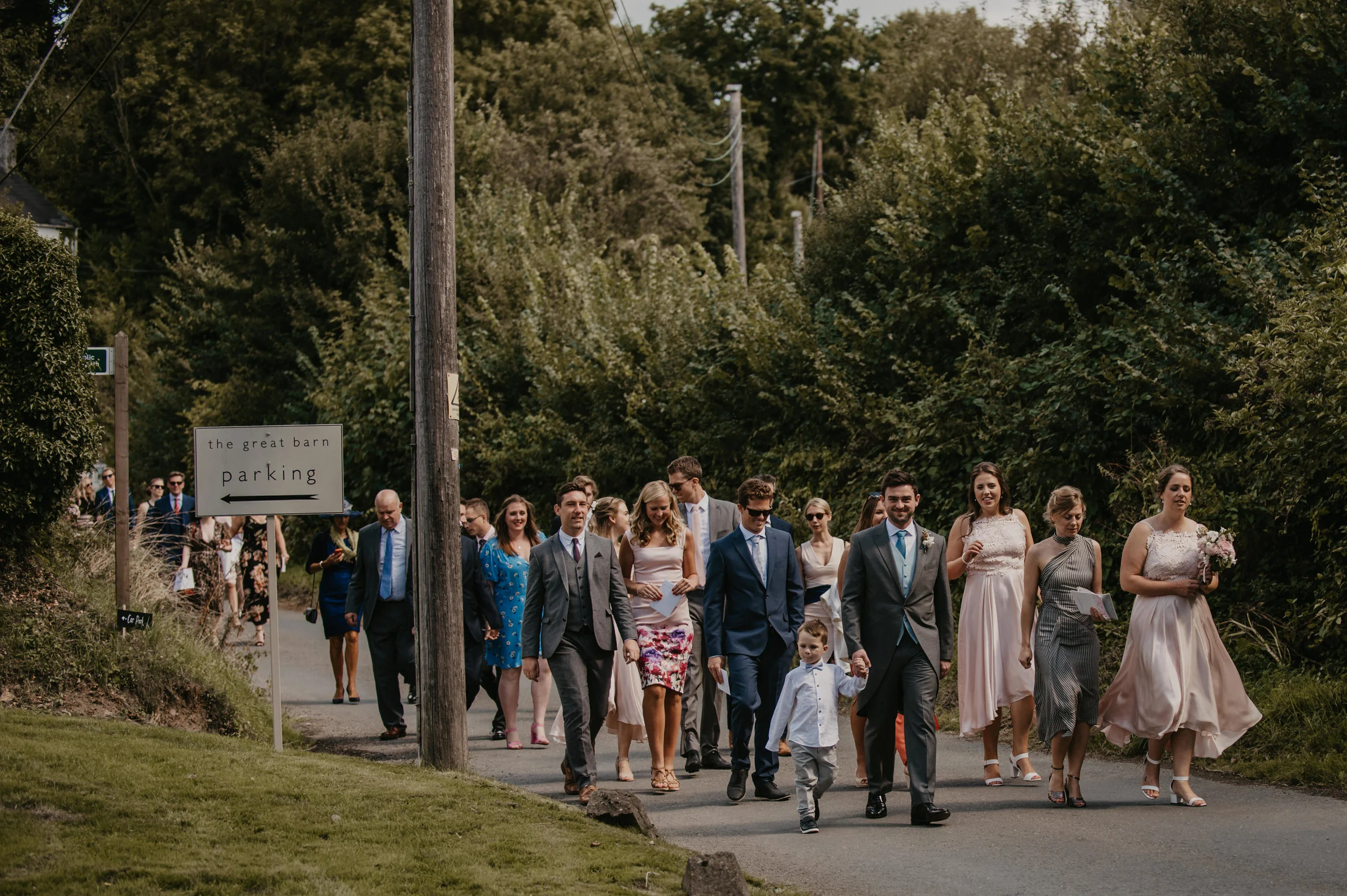 Group of people in formal attire walking outdoors, possibly at a wedding or celebration, with trees in the background and a sign pointing to parking