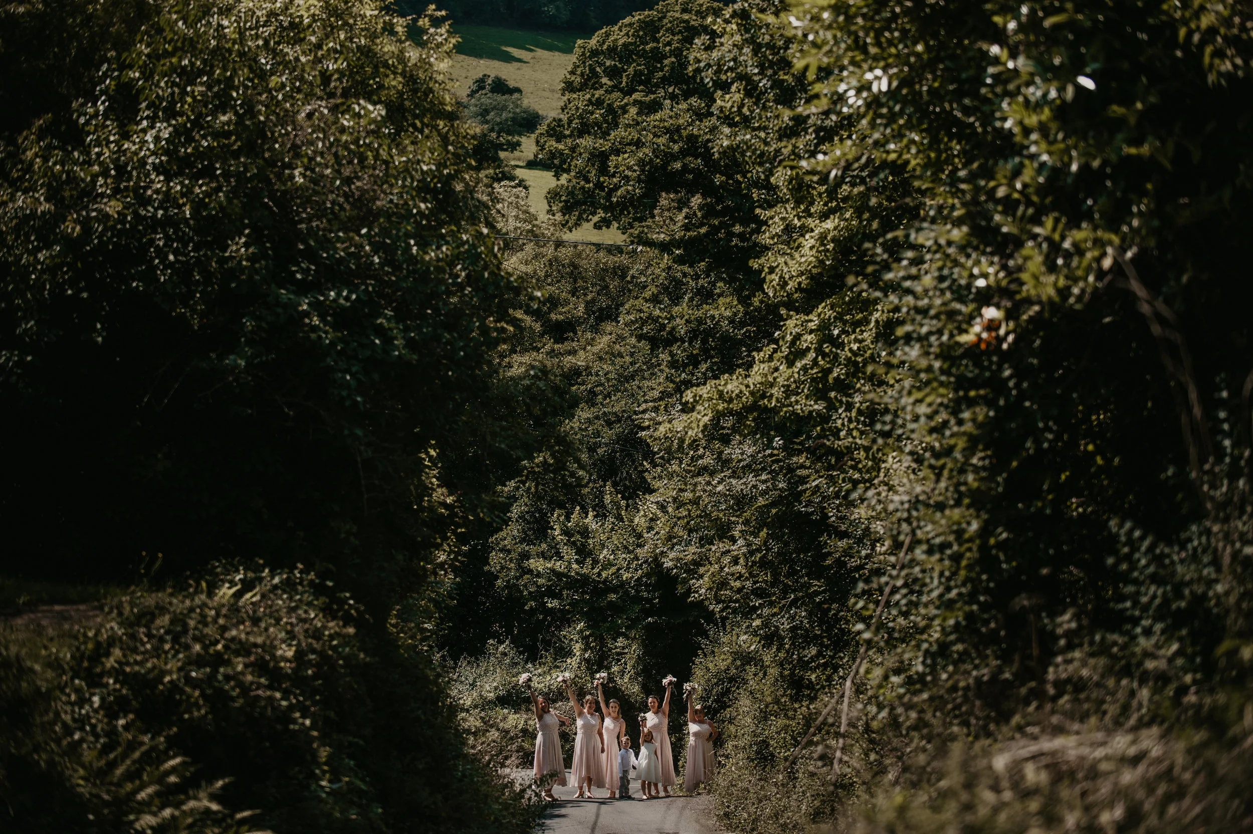 A group of wedding party members including bridesmaids and children standing on a rural road surrounded by dense green trees, some holding bouquets, with hills in the background on a sunny day.