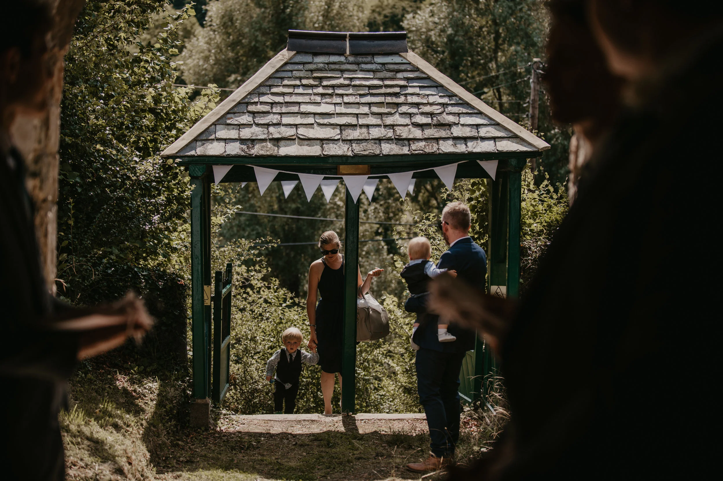 Group of people, including adults and children, entering a small pavilion or gazebo decorated with bunting, outdoors surrounded by trees and greenery.