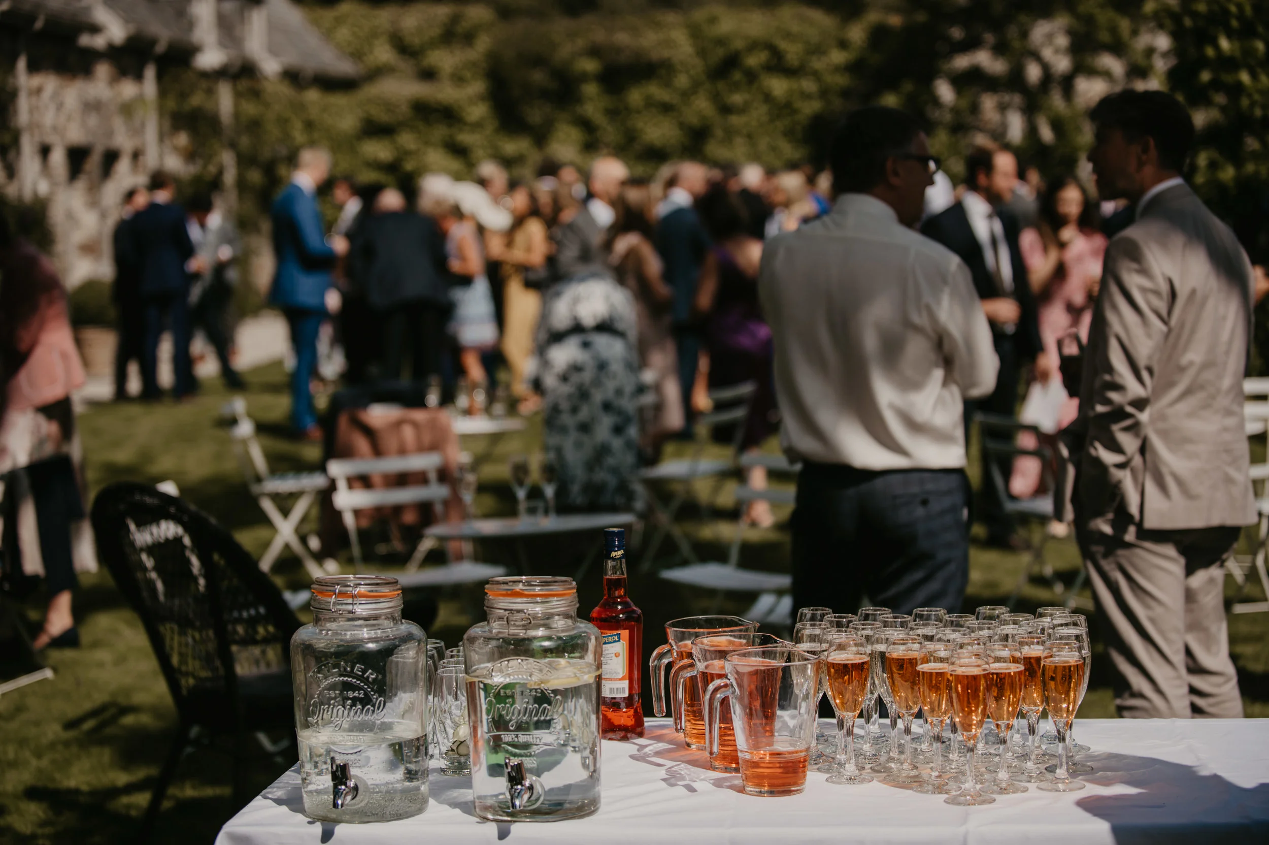 Table with pitchers of water, glasses of rosé wine, and a bottle of liquor at an outdoor gathering or wedding reception, with guests socializing in the background.