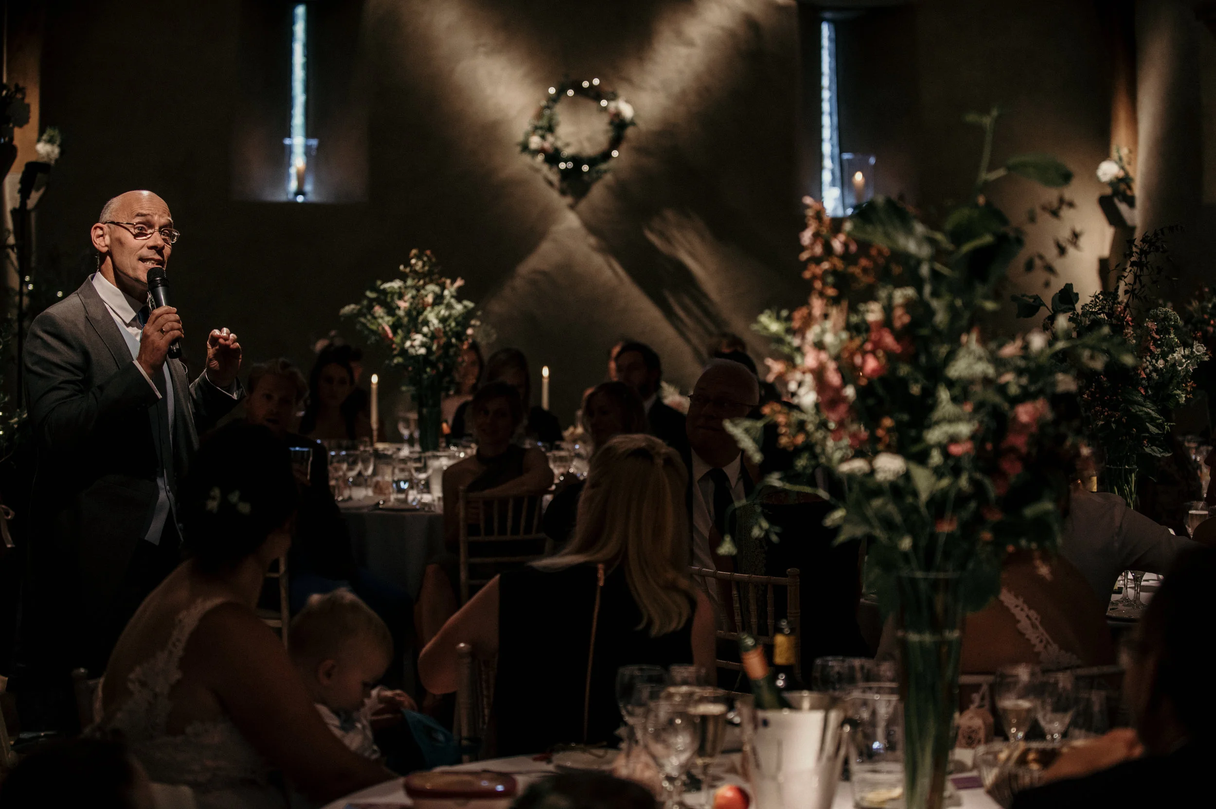 A man in a suit holding a microphone speaks at a formal event or wedding reception, with guests seated at decorated tables in a dim, elegant setting.