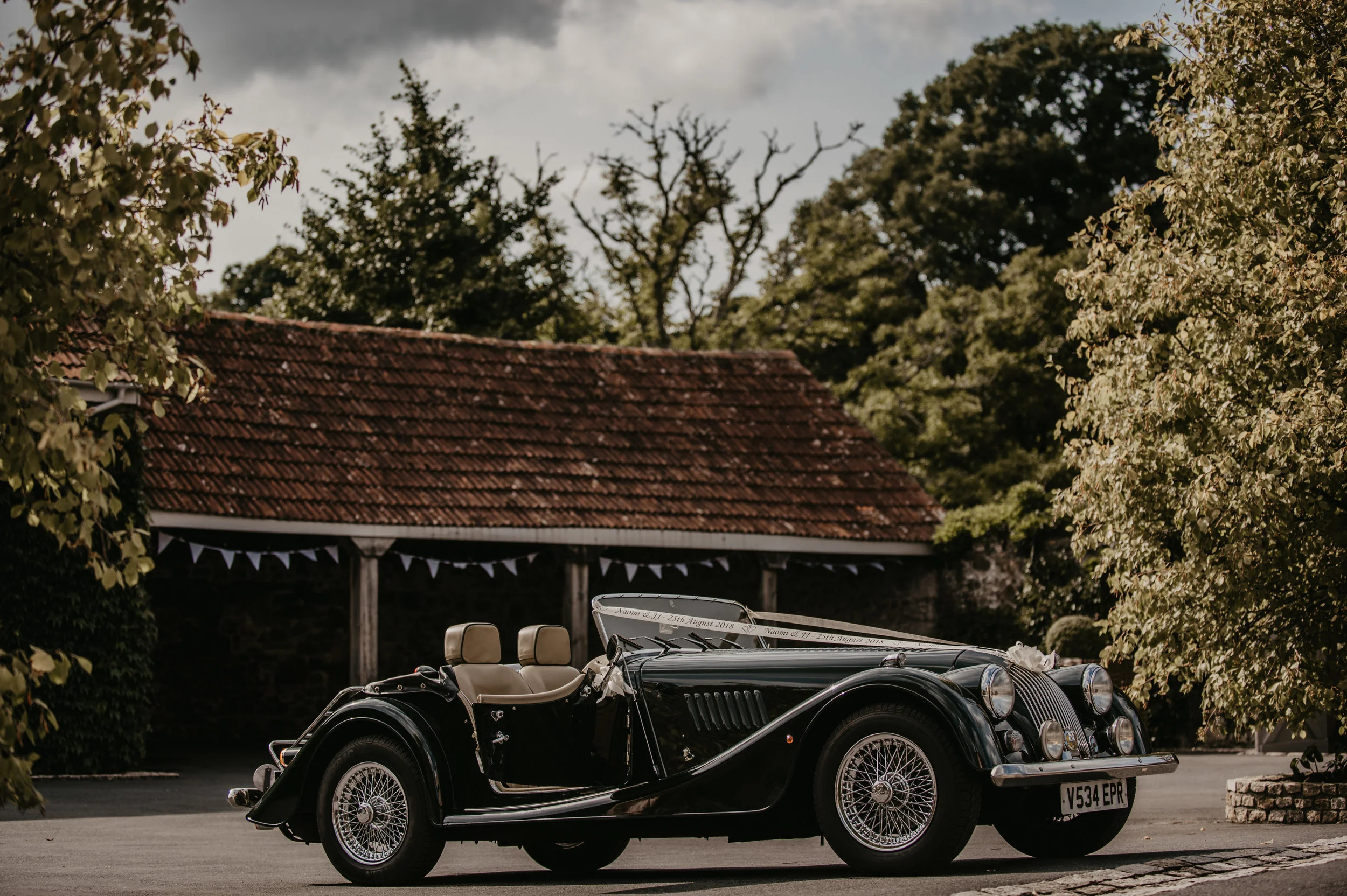 A vintage black convertible car parked on a paved driveway with trees and a building with a red-tiled roof in the background.