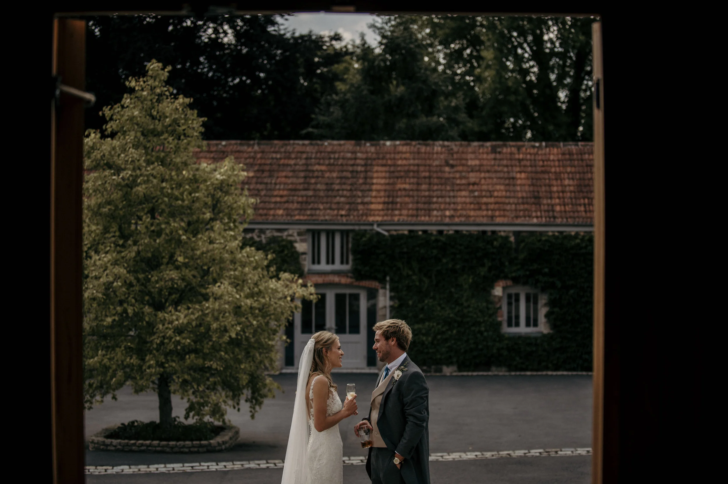 A bride and groom standing outdoors, holding glasses of champagne and looking at each other, framed by a doorway, with a tree, a house with ivy, and a paved area in the background.