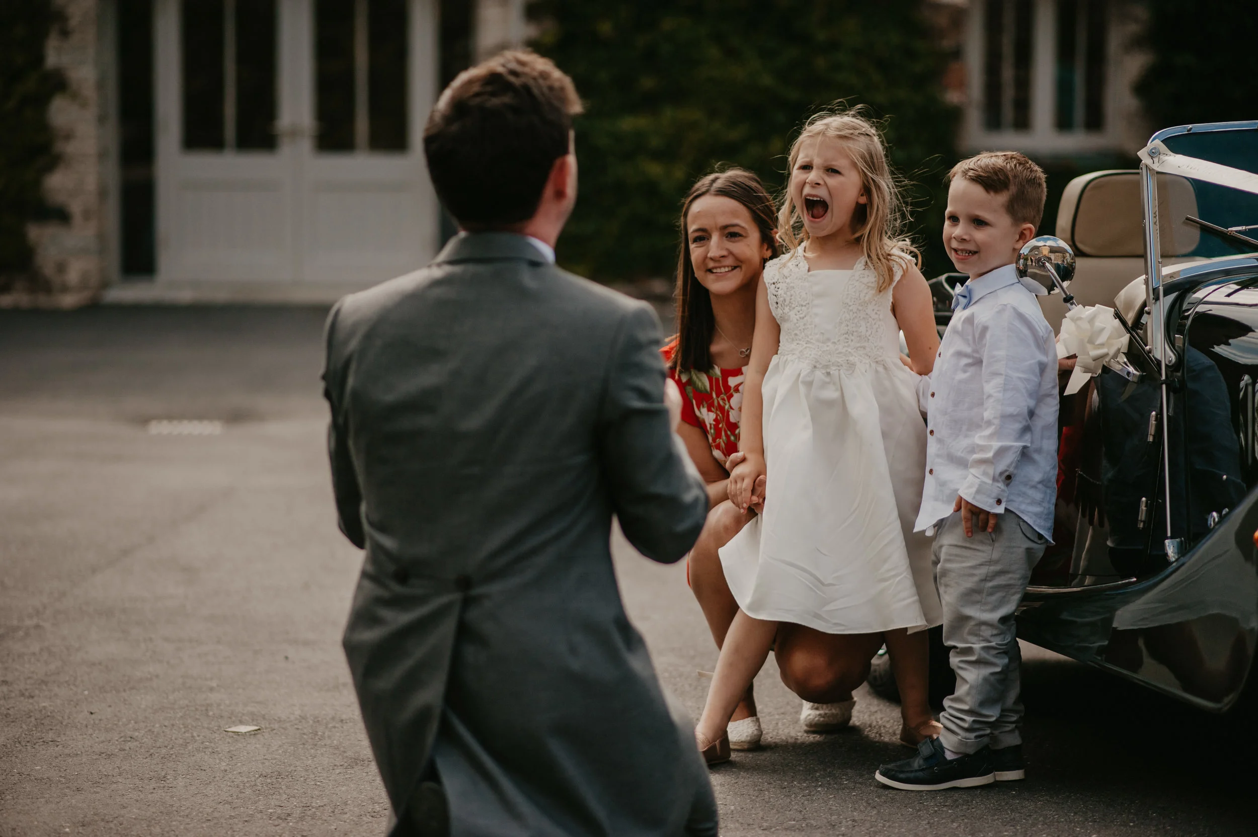 A man in a gray suit kneeling and holding hands with a young girl in a white dress, who is laughing, while a smiling woman and a young boy stand nearby near a vintage car outside.