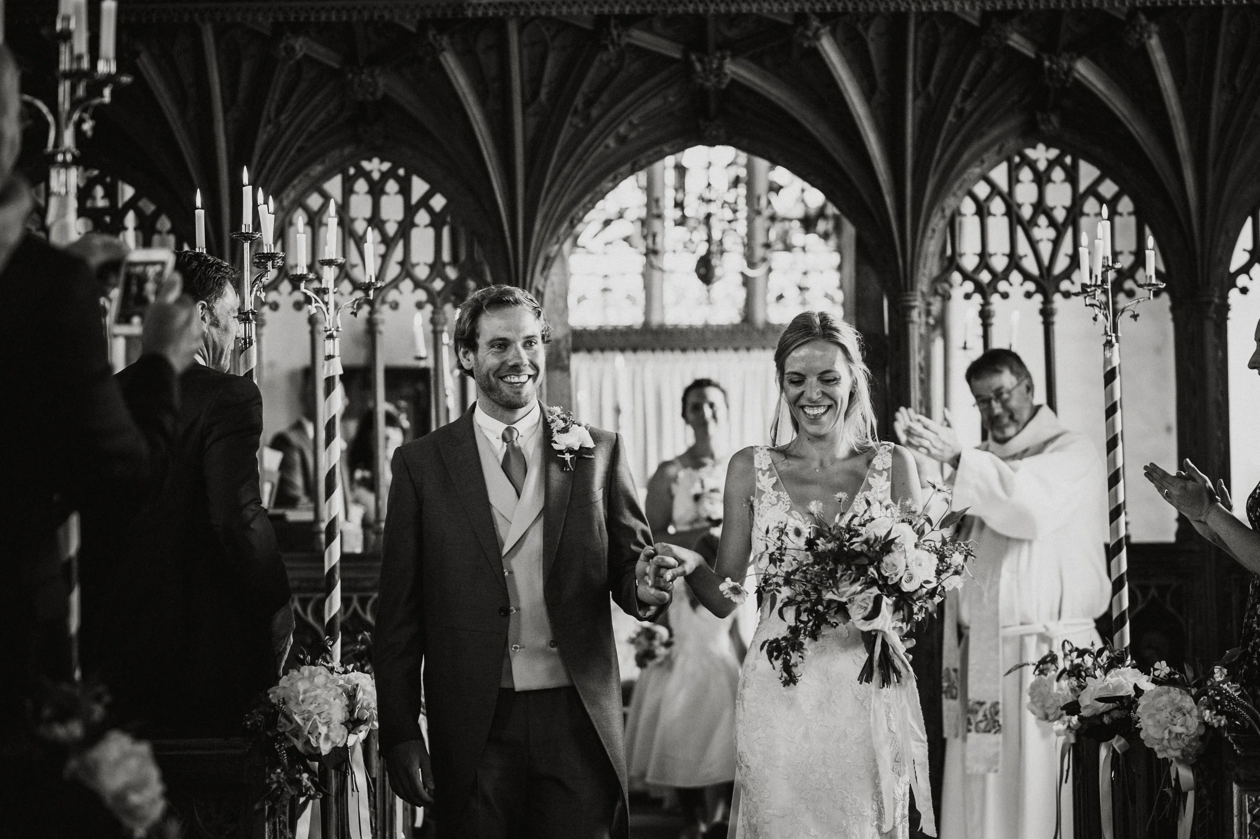 A black and white photo of a wedding ceremony with a bride and groom smiling and holding hands, walking down the aisle in a church with Gothic architectural elements. The bride is holding a bouquet and wearing a lace dress, while the groom is in a su