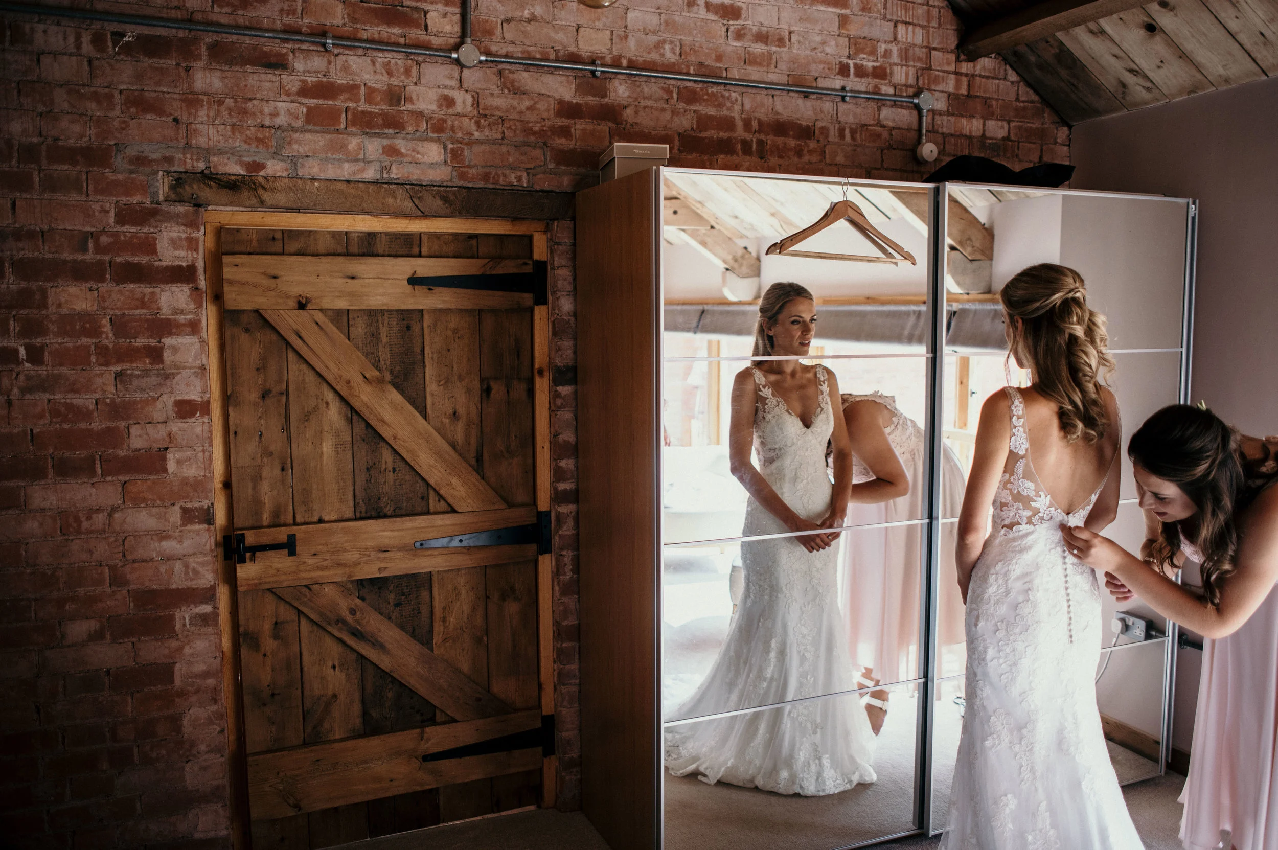 Two women in wedding dresses are getting ready, one standing and looking at herself in a mirror while the other is adjusting her dress. They are in a rustic room with brick walls and a wooden ceiling.