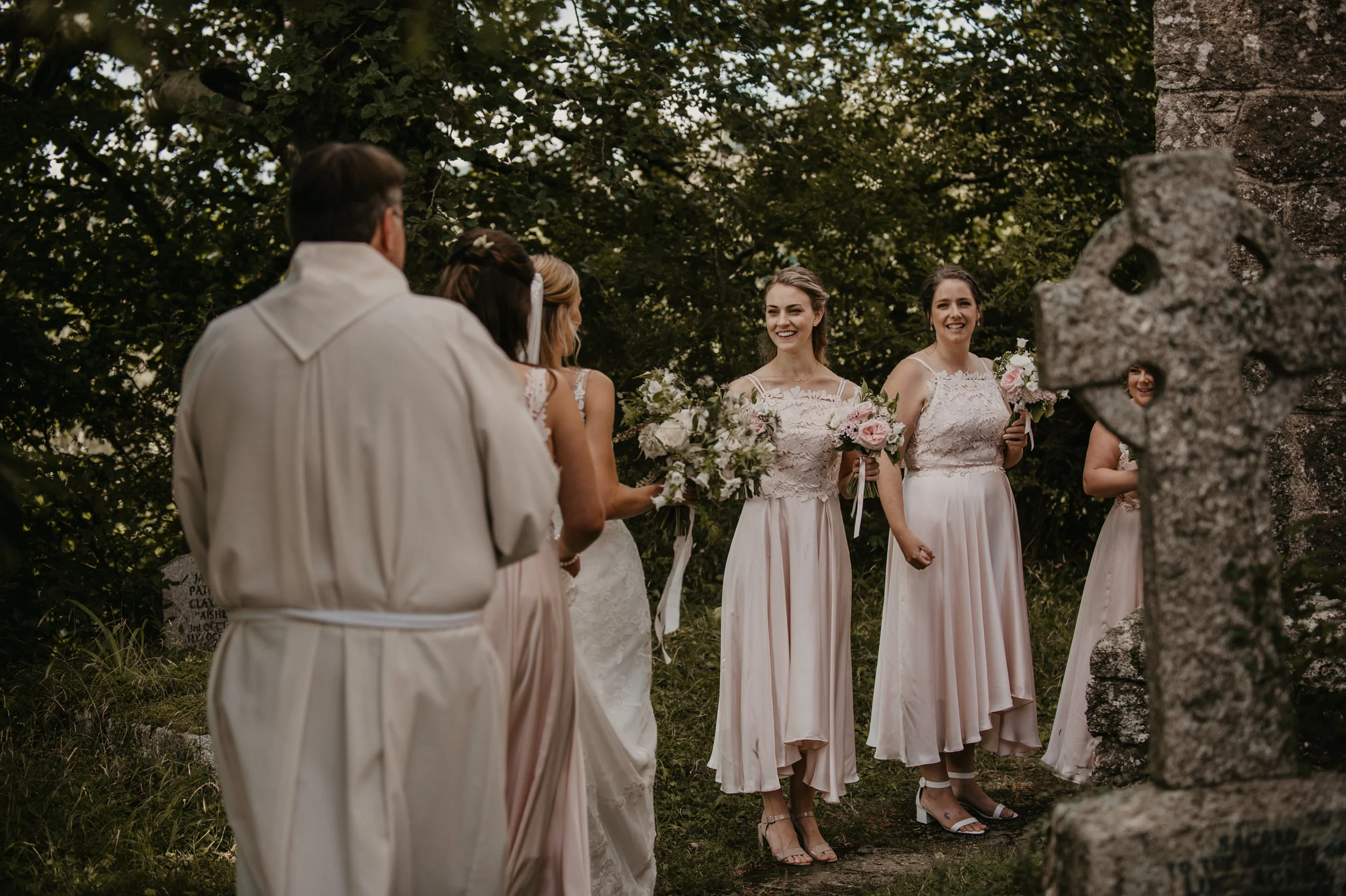 A wedding ceremony outdoors with a group of women in light pink dresses and a man in a light-colored suit, with trees and a cross-shaped stone monument in the background.