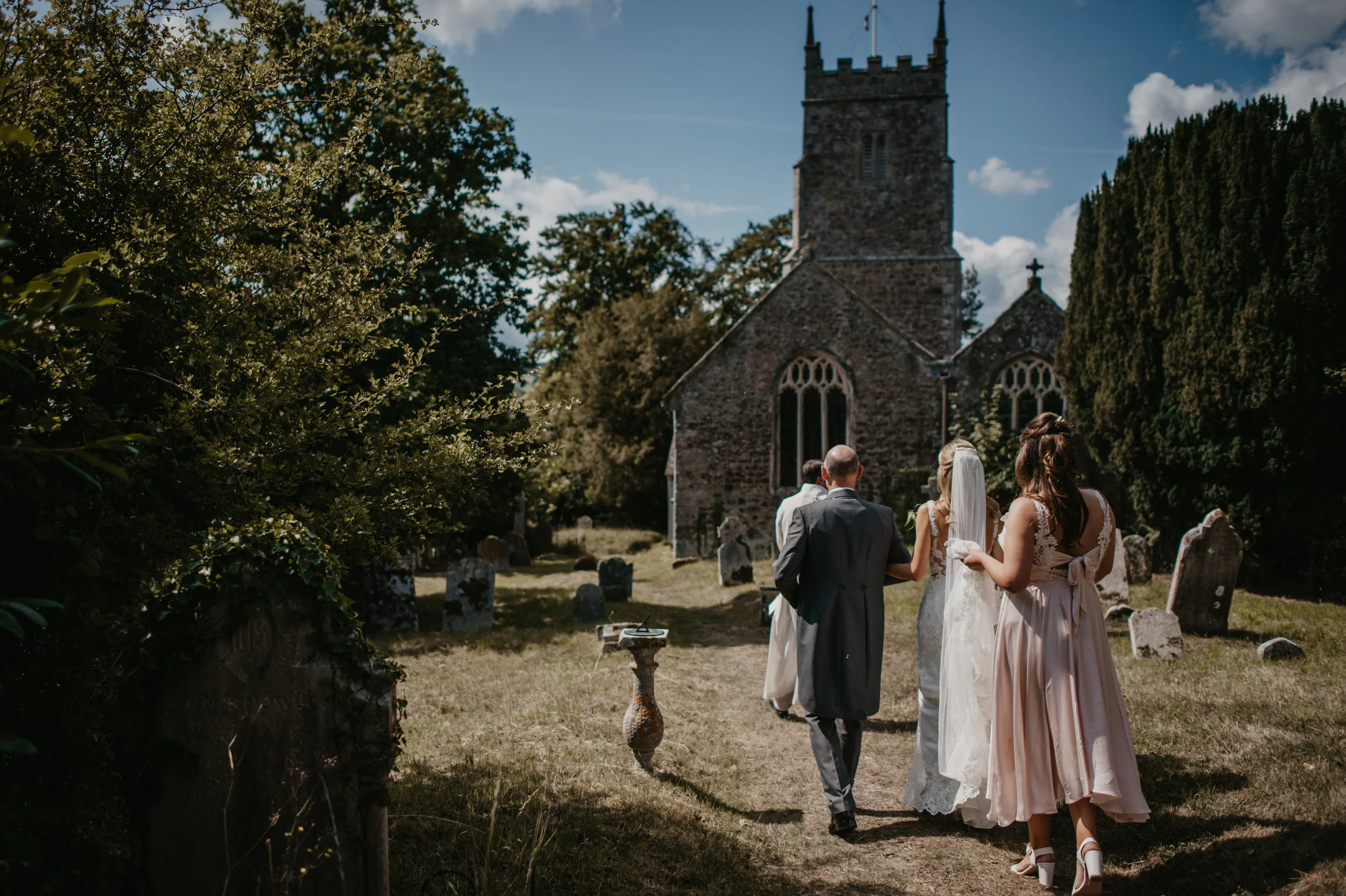 A wedding procession walking toward a church on a sunny day with blue skies, surrounded by greenery and tombstones.