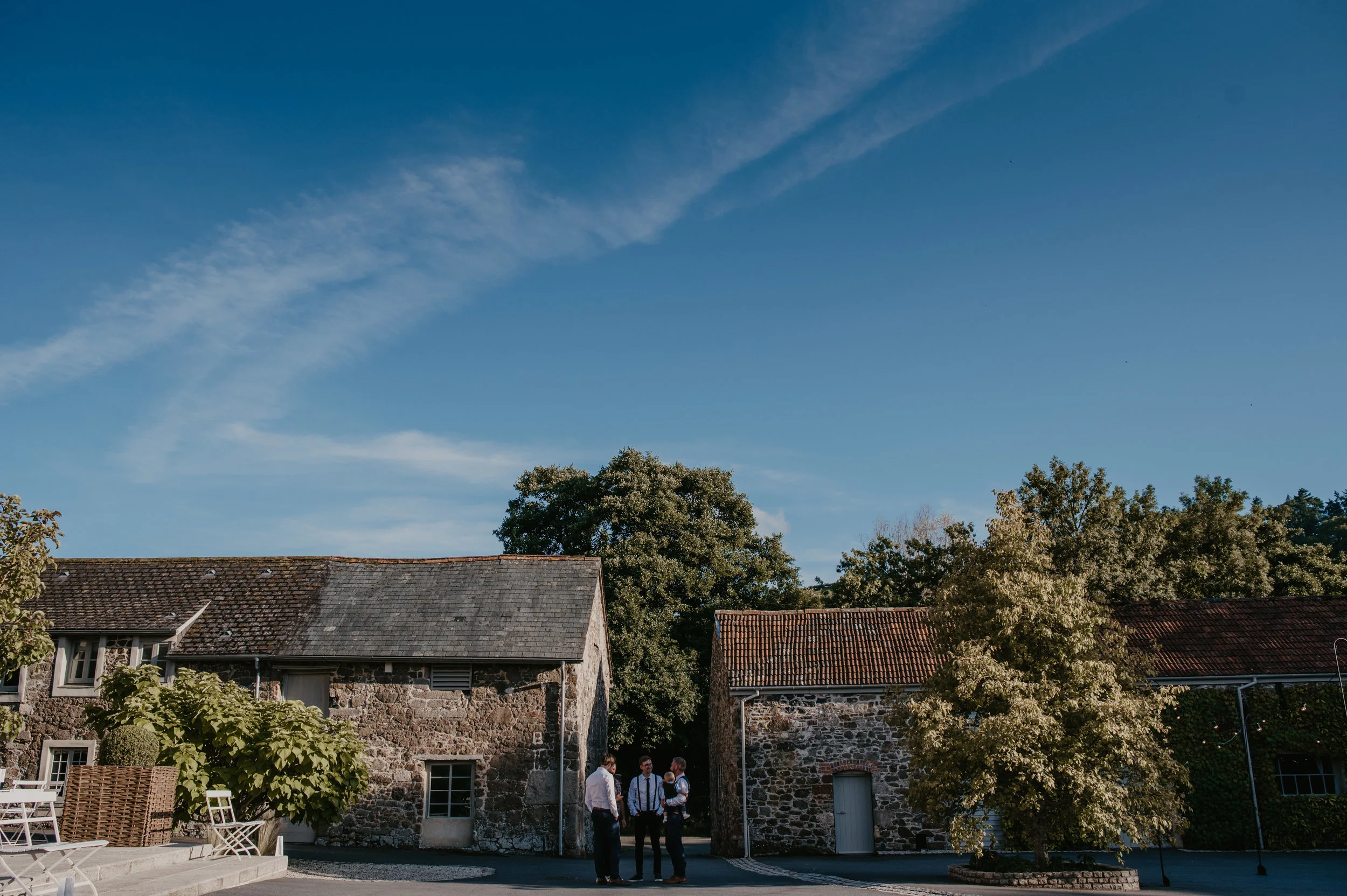 Three men stand outside between two old stone buildings, under a blue sky with some clouds, in a rustic outdoor setting.