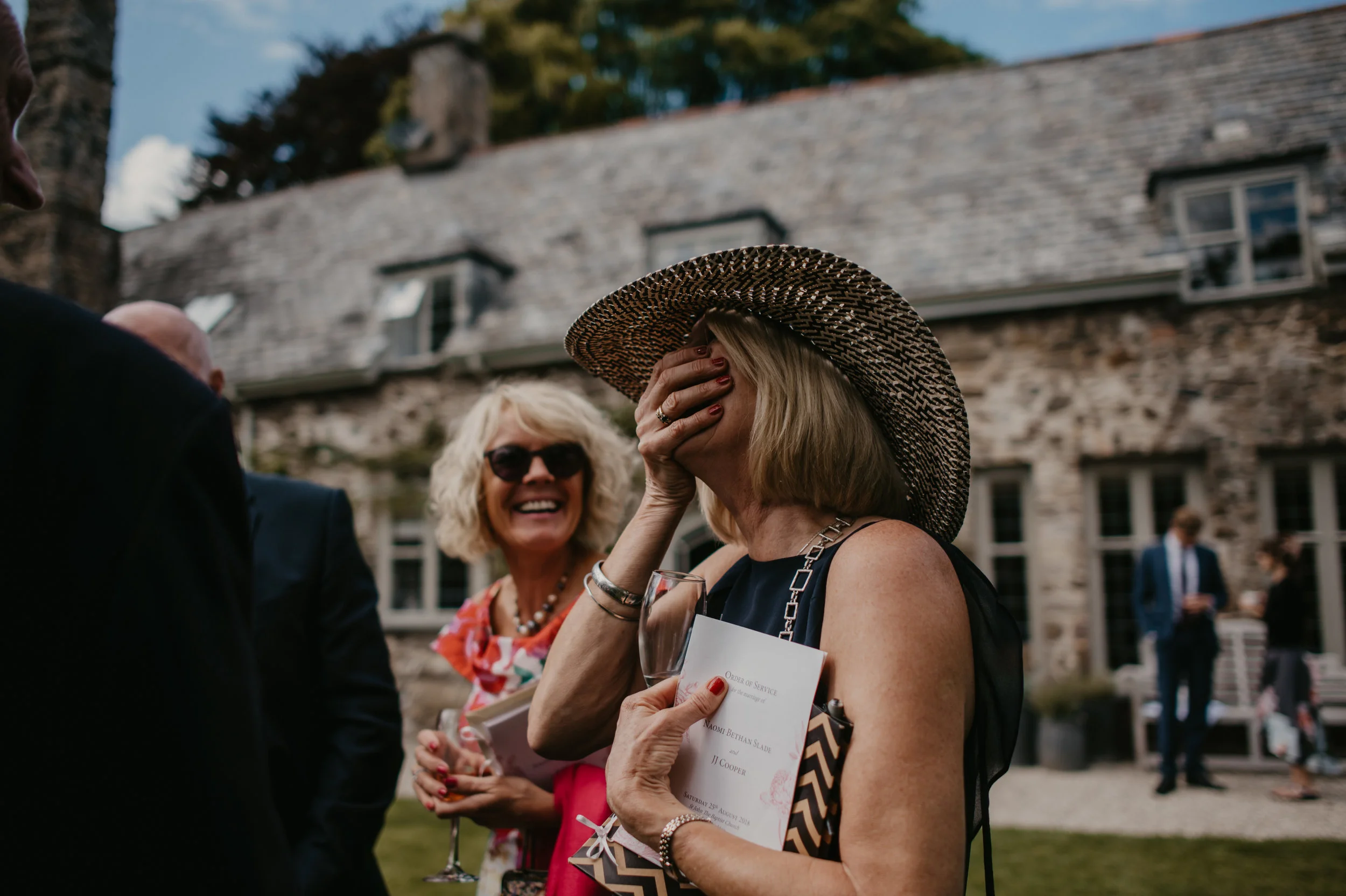 A woman in a large sun hat covering her face with her hand, holding a glass of wine and a ceremony program, standing outdoors in front of an old stone building during a wedding celebration. Another woman in sunglasses and a colorful dress is smiling 