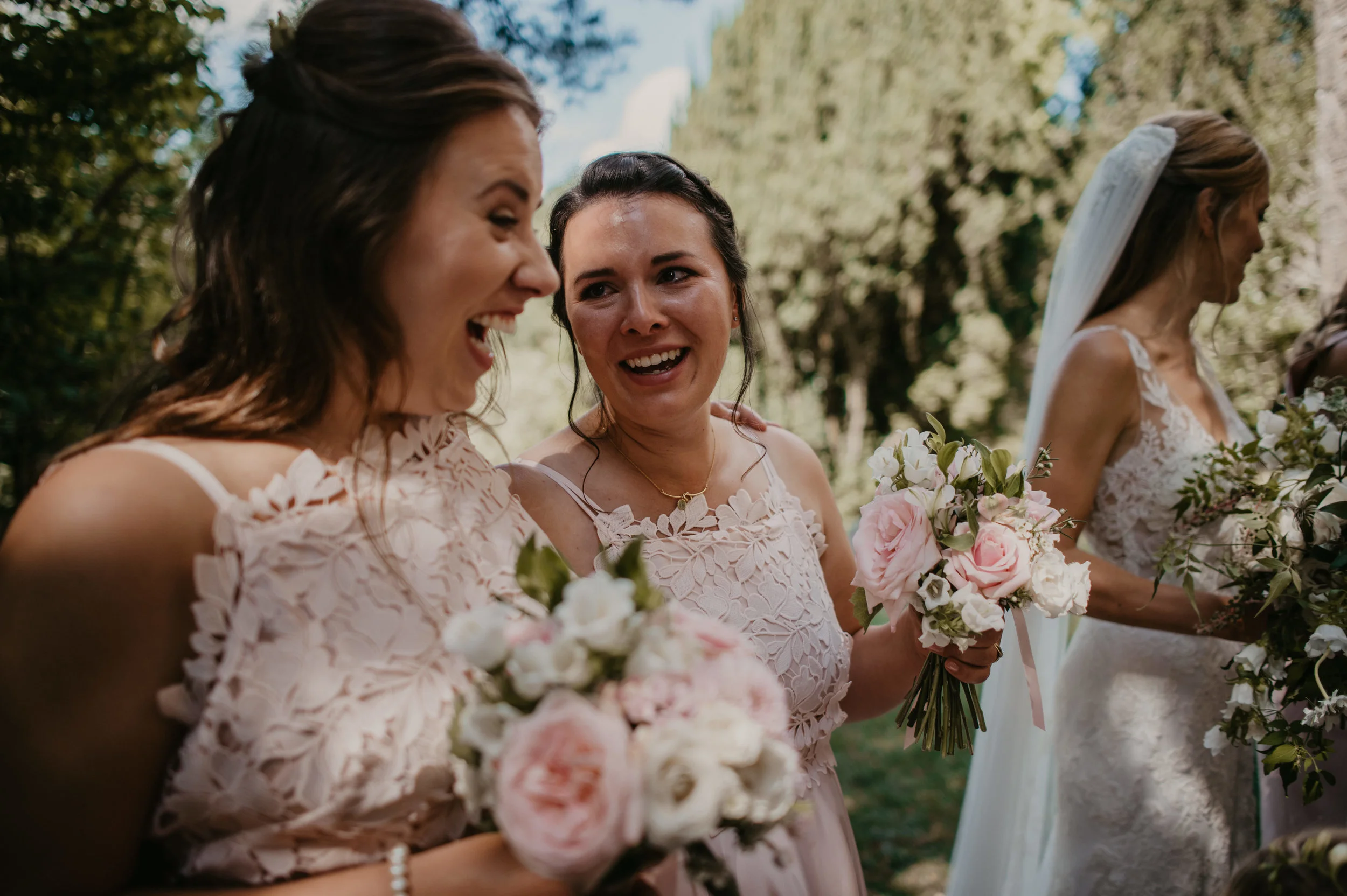 Four women in wedding dresses holding bouquets of pink and white flowers, standing outdoors under trees during a wedding celebration, with soft sunlight filtering through the leaves.