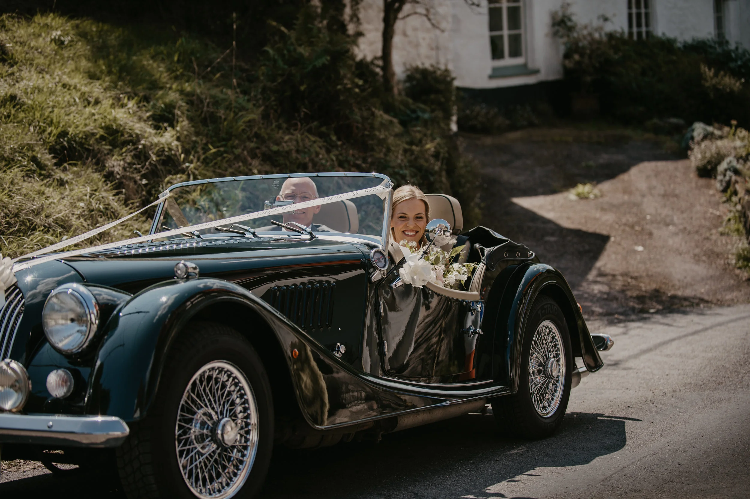 A bride and groom driving in a vintage black convertible car decorated with white flowers, smiling and enjoying their wedding day, on a rural road with greenery and a house in the background.