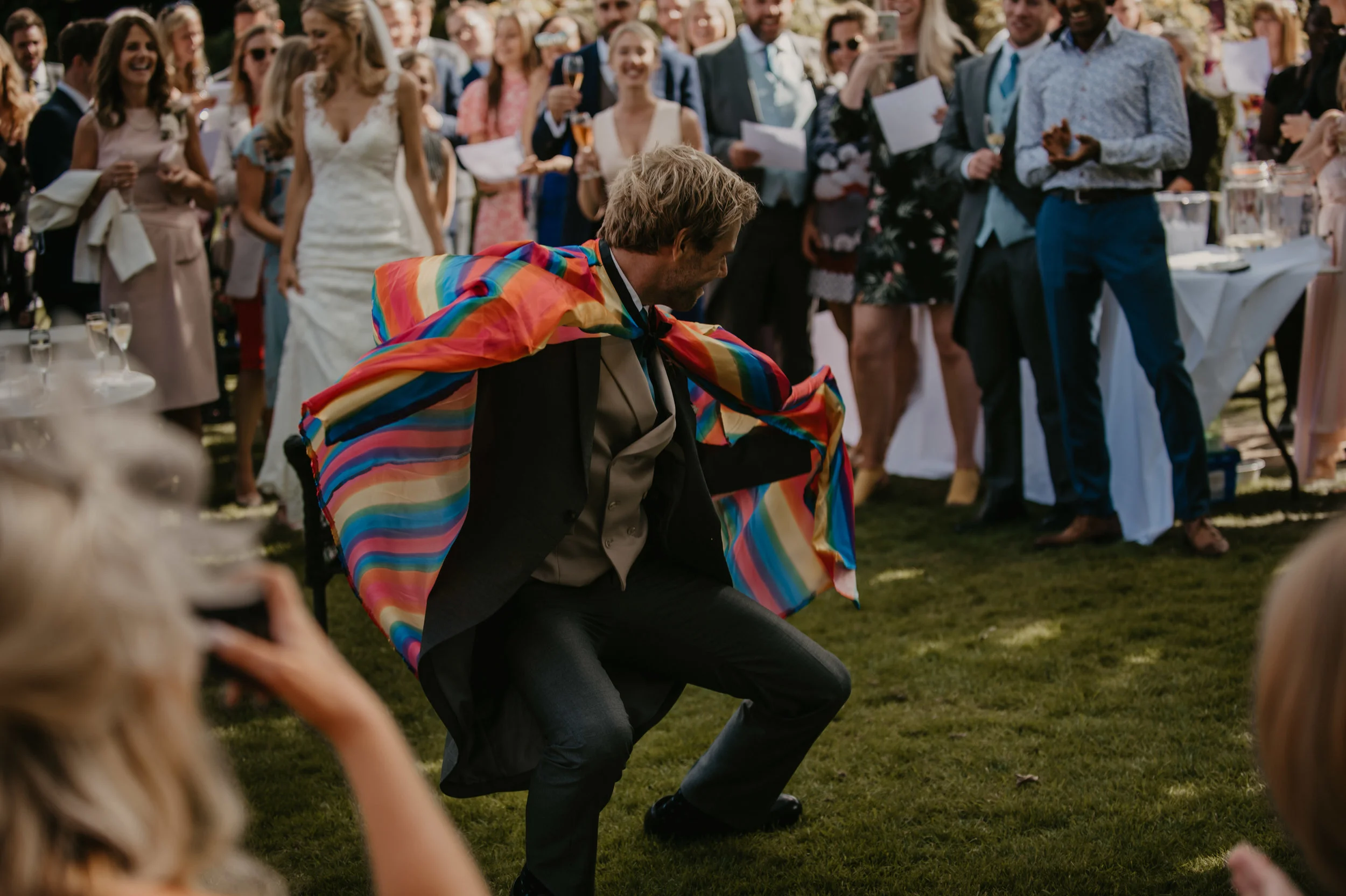 A man in a tuxedo dancing at a wedding reception, draped with a rainbow-colored cloth, while guests watch and cheer around him.