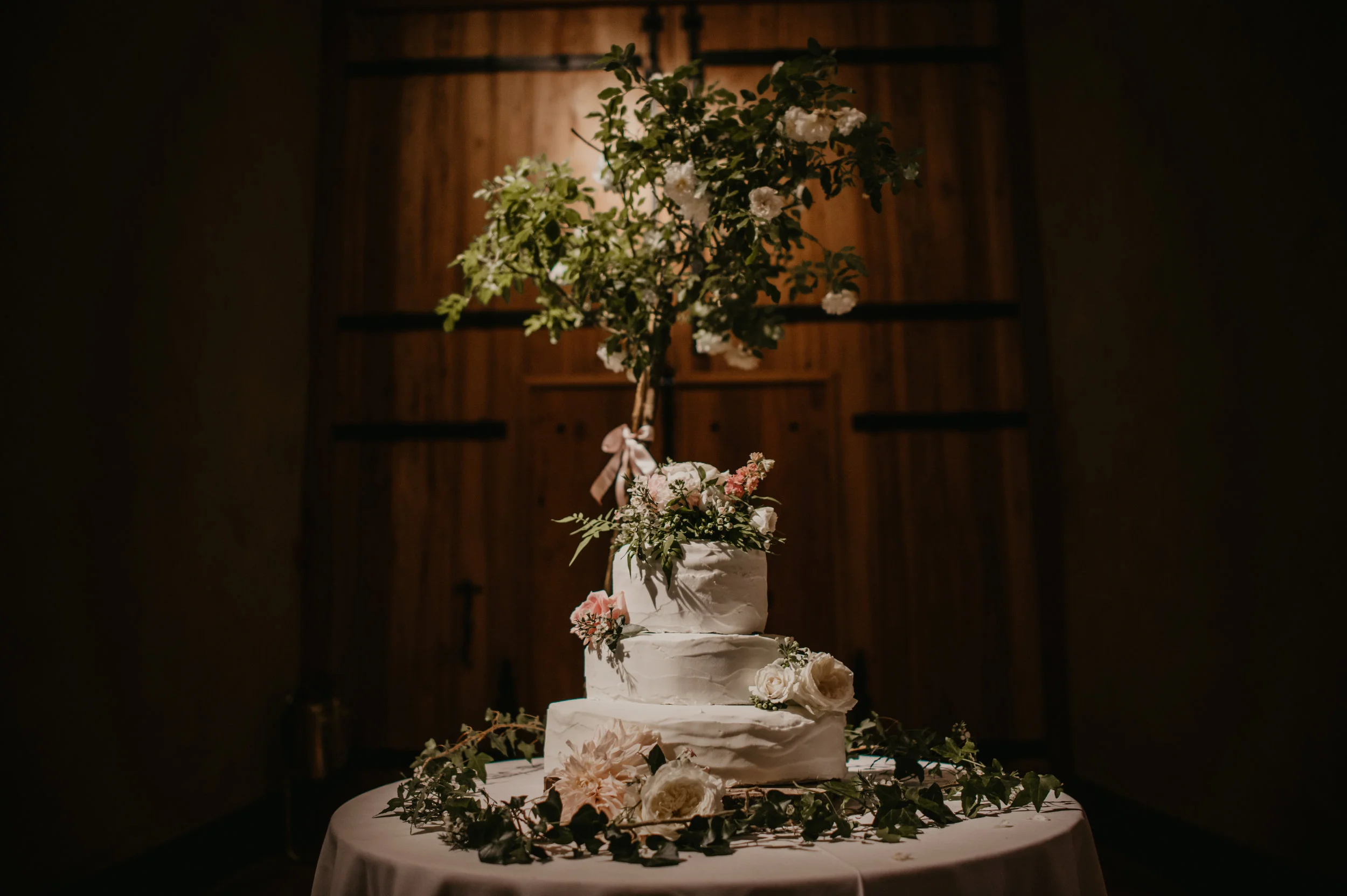 A three-tier wedding cake decorated with white roses, pink flowers, and greenery on a table draped with a white cloth, with a large floral arrangement on top, all inside a dimly lit room with wooden walls.