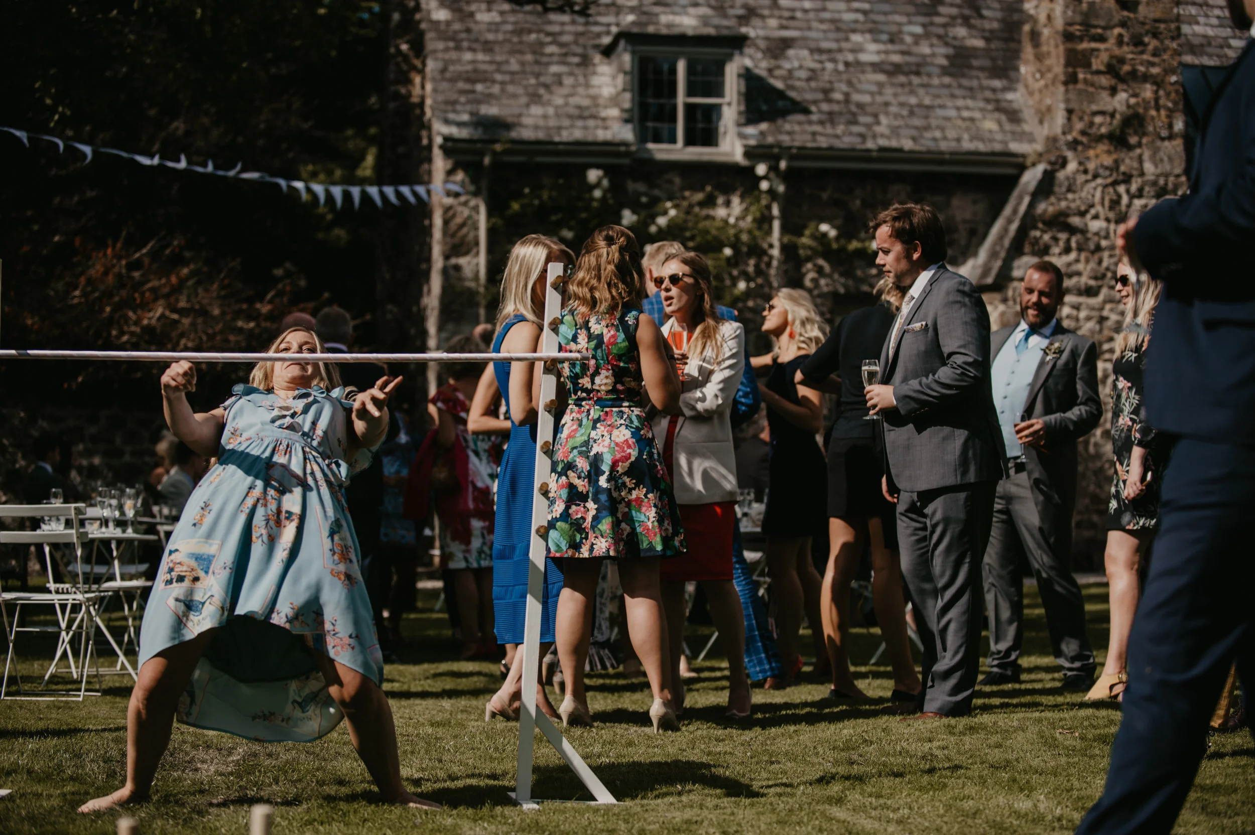People at an outdoor wedding reception playing a limbo game on the grass, with a woman in a floral blue dress holding her position under a limbo stick. Guests are dressed in semi-formal and formal attire, some holding drinks, with a stone building in