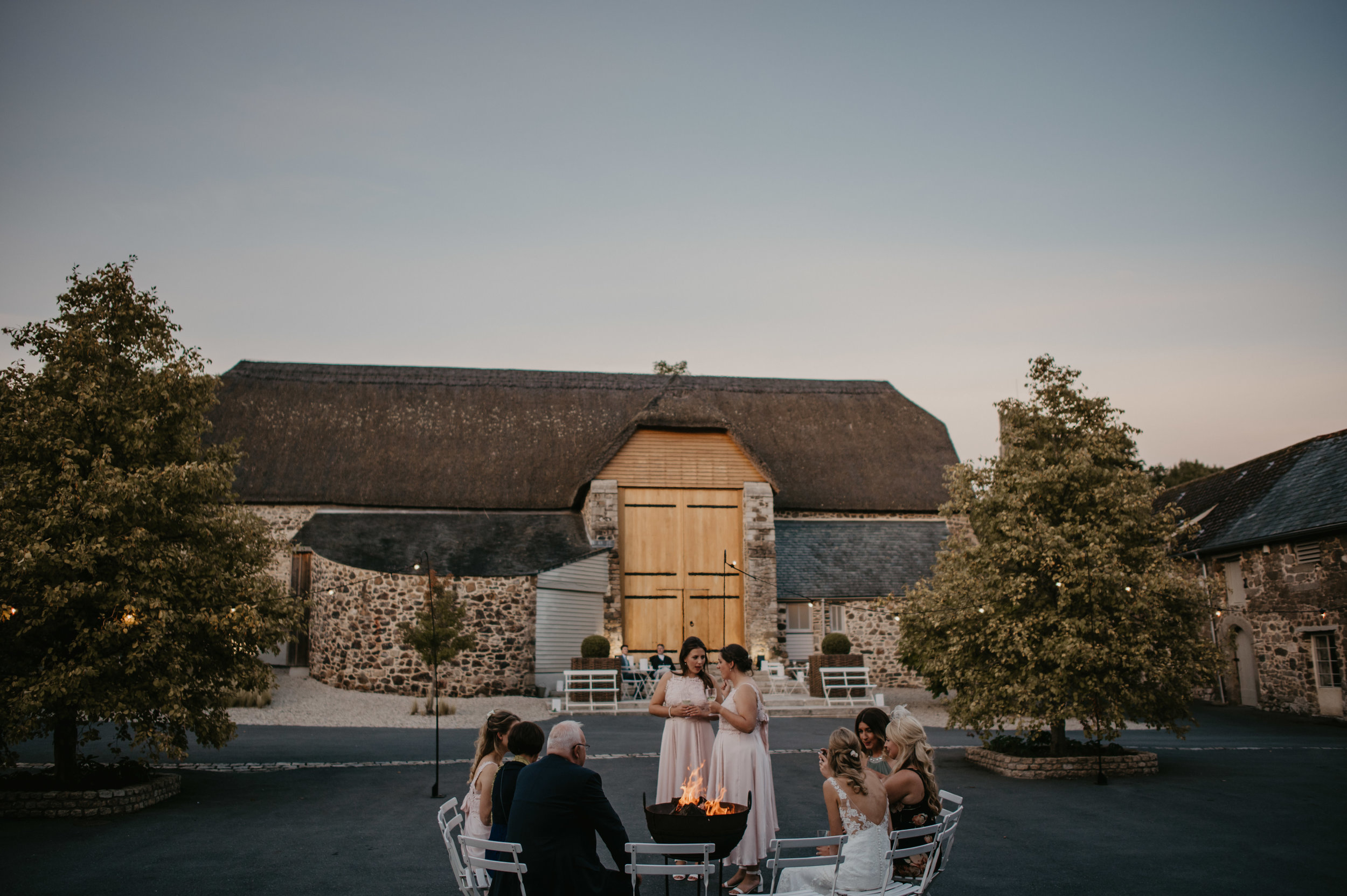 A wedding reception outdoors during dusk, with seven people sitting around a fire pit, two women standing and talking, and a rustic barn in the background.