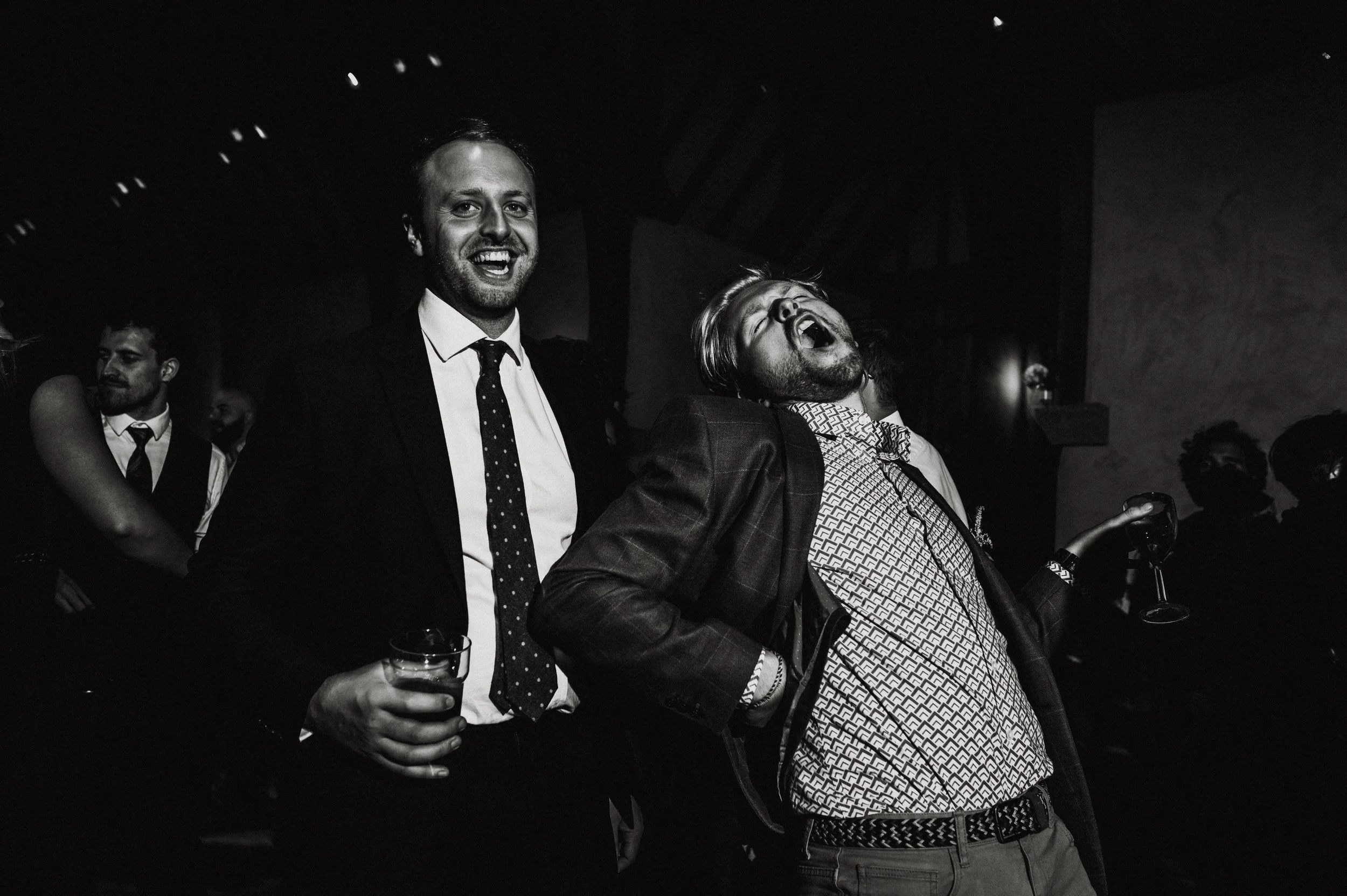 Black and white photo of two men at a party, one holding a glass of drink and smiling, the other laughing with head thrown back and holding a glass of wine, with more people in the background.