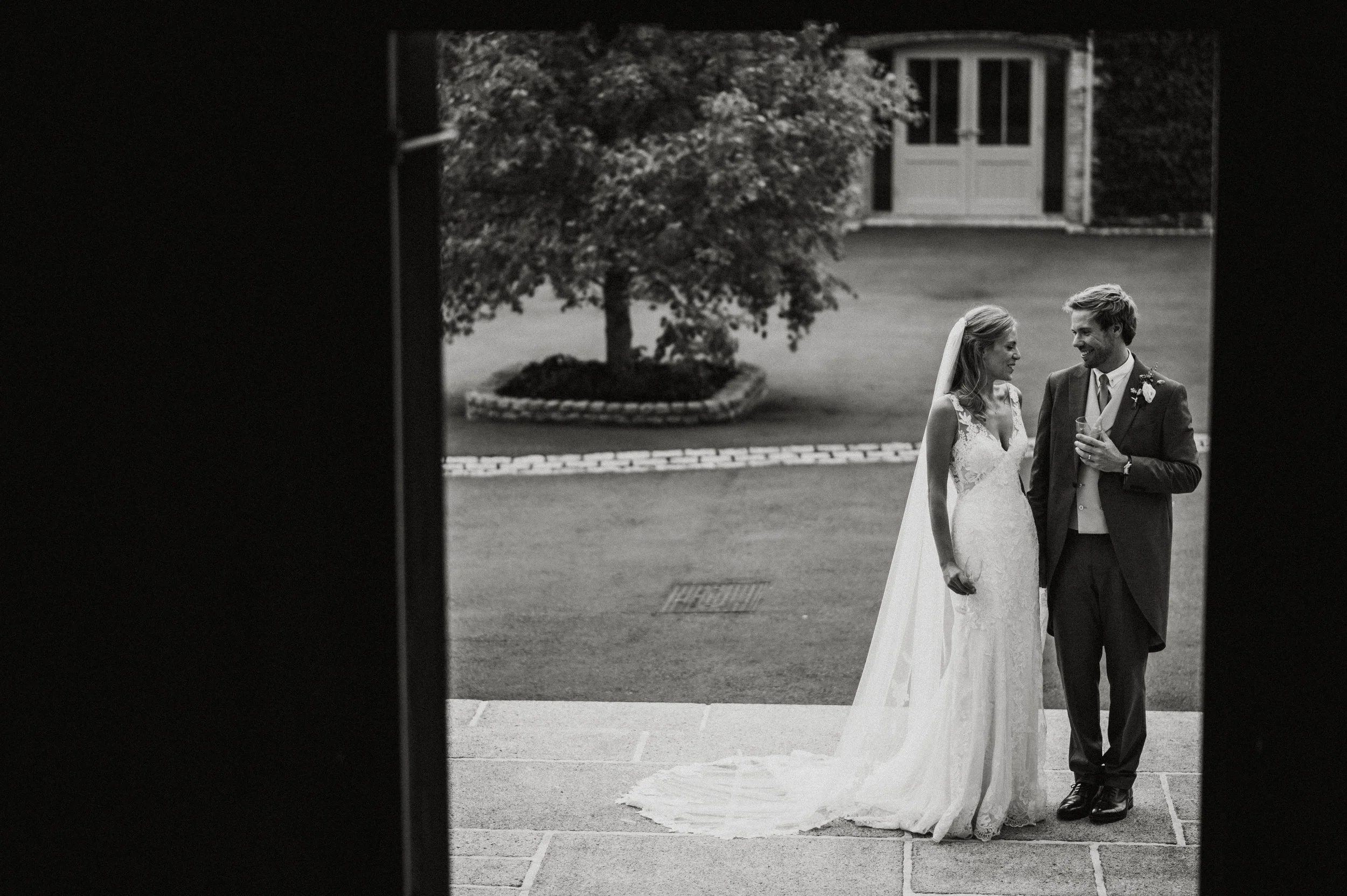 A black-and-white photograph of a bride and groom standing outdoors, sharing a moment. The bride wears a lace wedding gown with a long veil, and the groom wears a suit with a boutonniere, holding a drink. They are smiling at each other, with a tree a