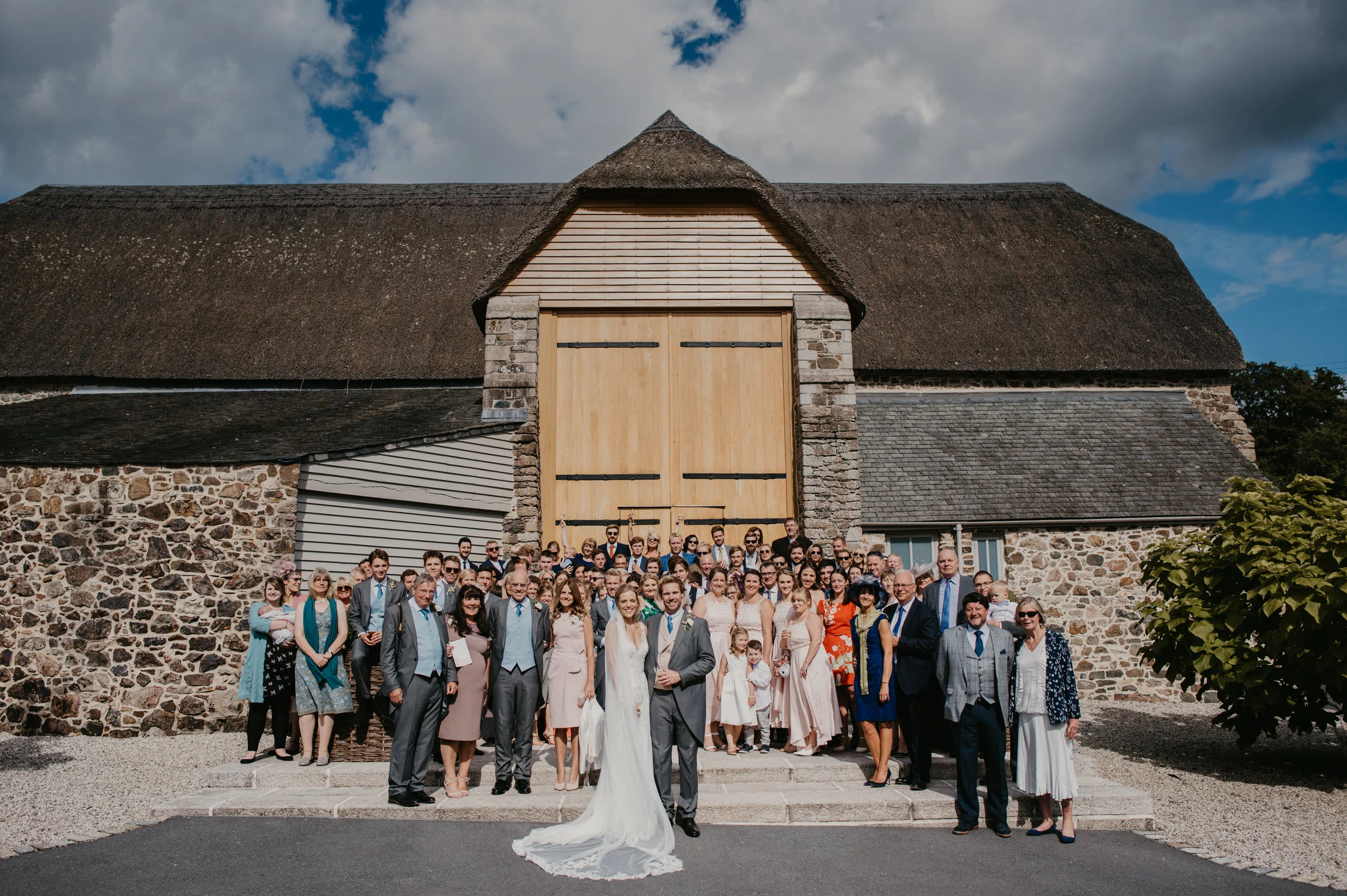 A large group of people dressed in formal attire gathering outside a rustic stone and wooden barn for a wedding celebration. The bride and groom stand in front, smiling.