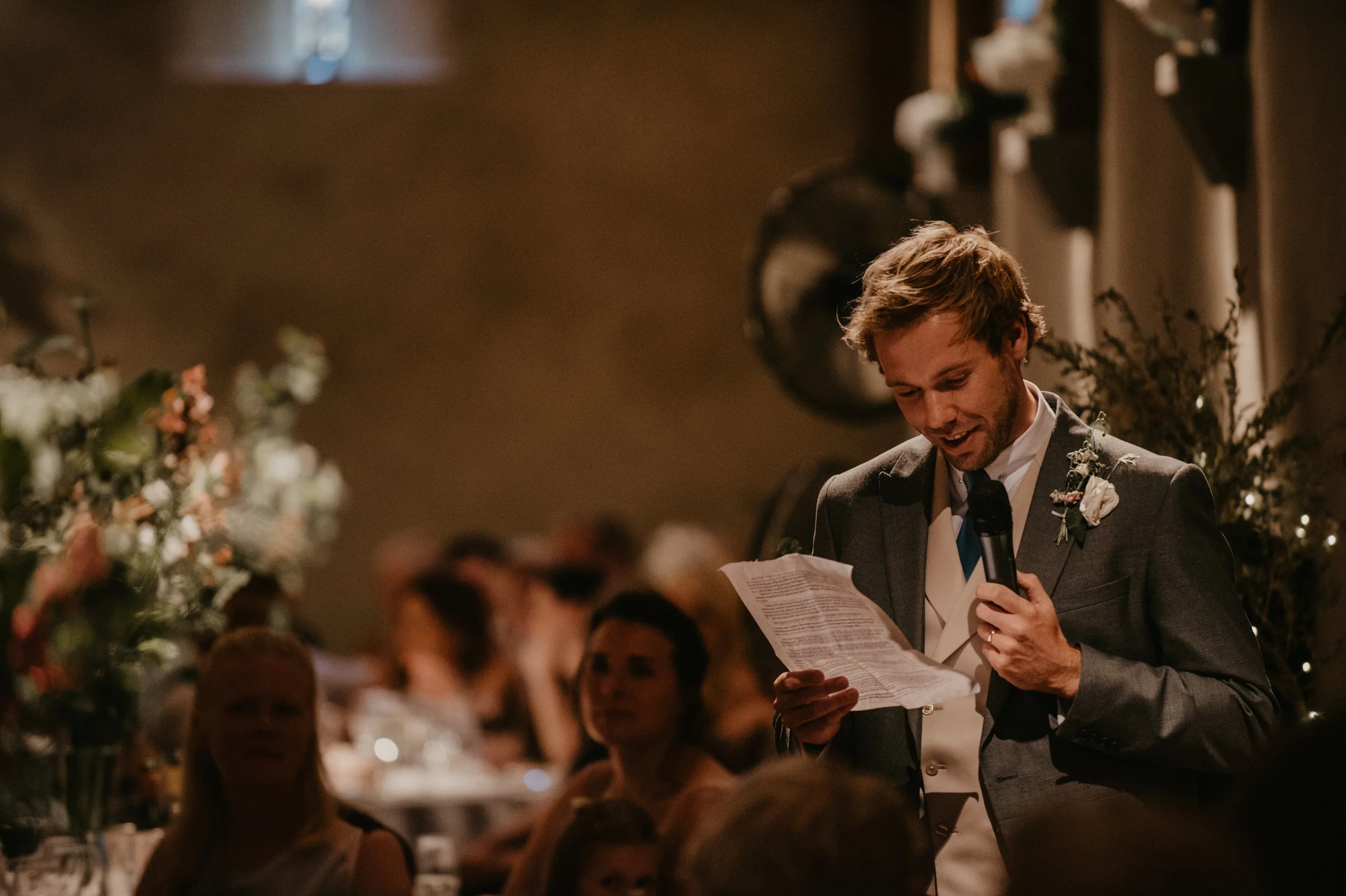 A man in a suit giving a speech at a wedding reception, holding a microphone and reading from a paper, with guests seated at tables in the background.