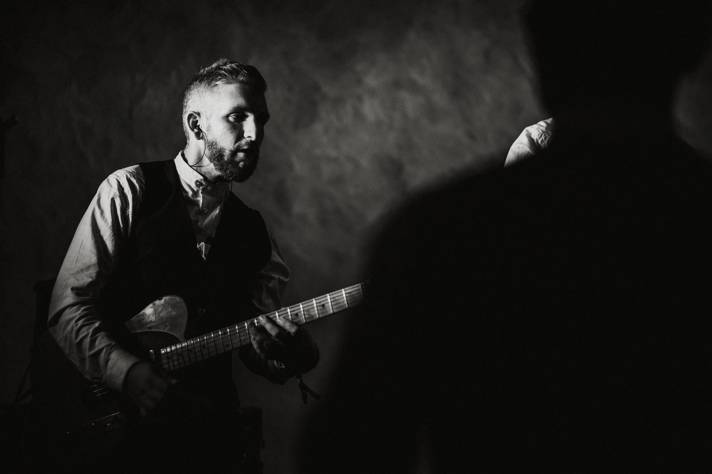 A black and white photo of a man with a beard playing an electric guitar, wearing a shirt and vest, with a serious expression, in a dimly lit room with an uneven wall background.