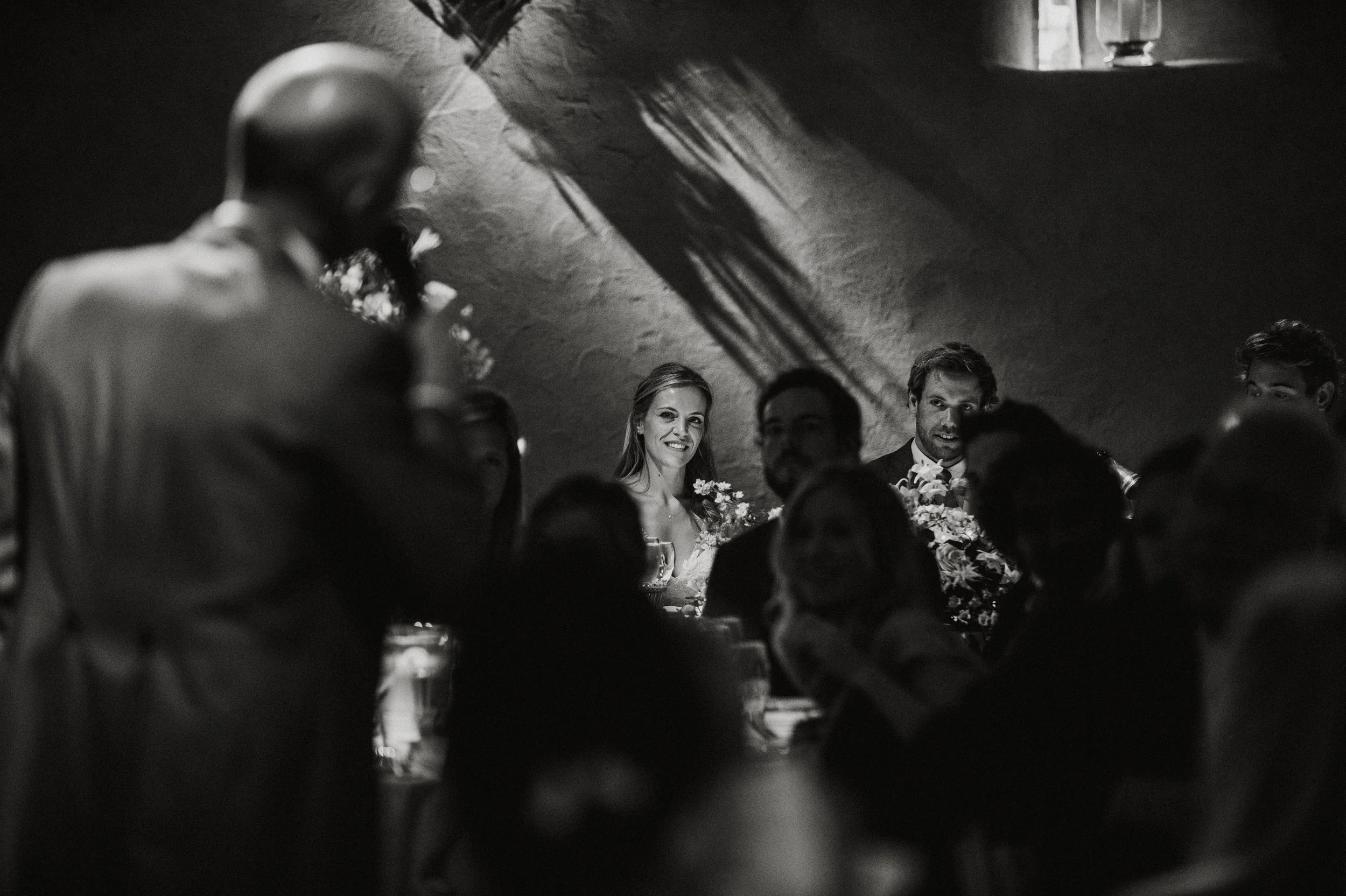 Black and white photo of a wedding reception with many guests sitting at a table, a woman and a man with flowers are in the center, and a man is standing on the left side of the image.