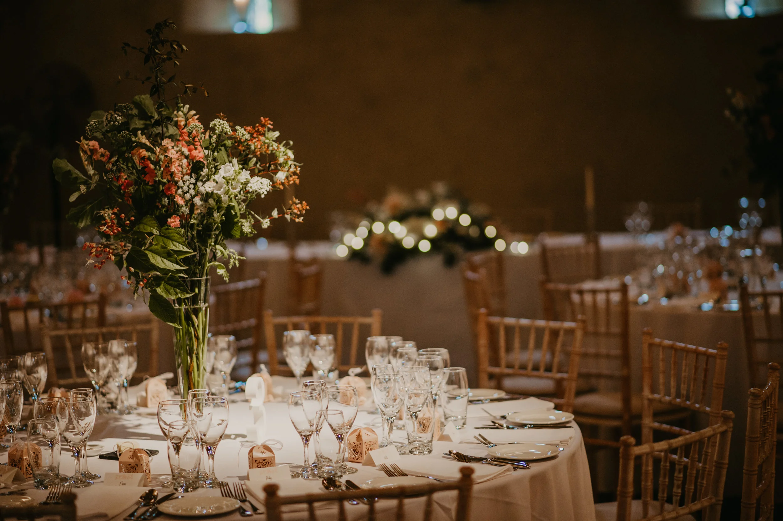 A decorated banquet table with a large floral centerpiece and arranged glassware, plates, and silverware in a dimly lit event space with string lights in the background.