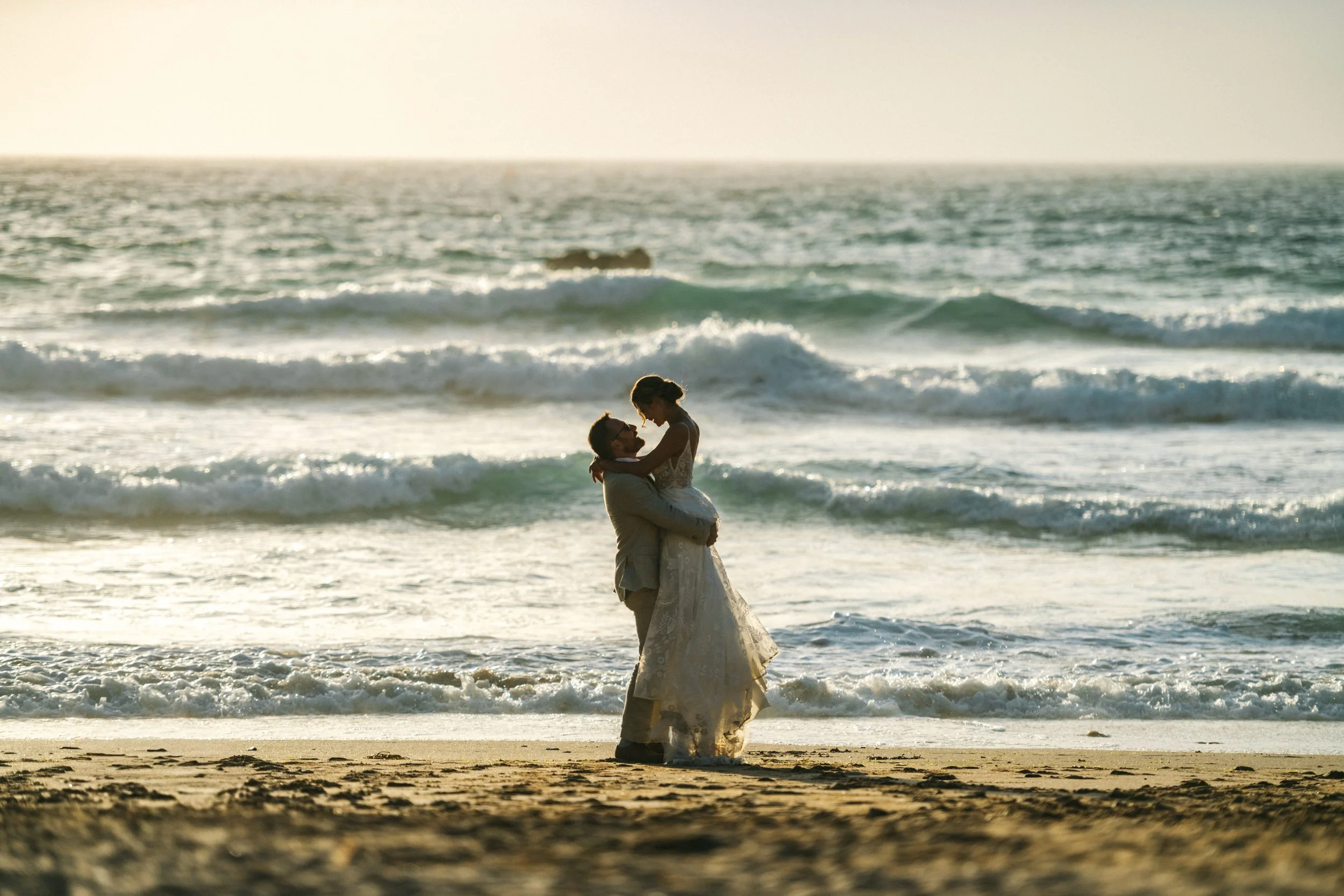Bride and groom embracing on a beach with ocean waves in the background at sunset.