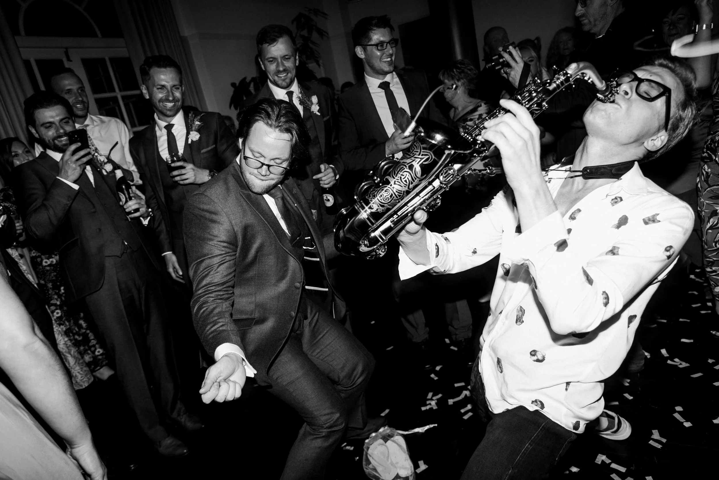 Black and white photo of a lively wedding party in Cornwall with a saxophonist playing energetically and a group of people dancing and enjoying the music.