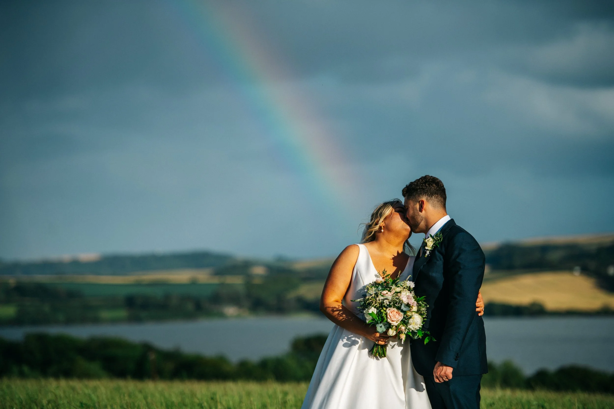 Bride and groom kissing under a rainbow in a scenic outdoor farm setting in Cornwall.