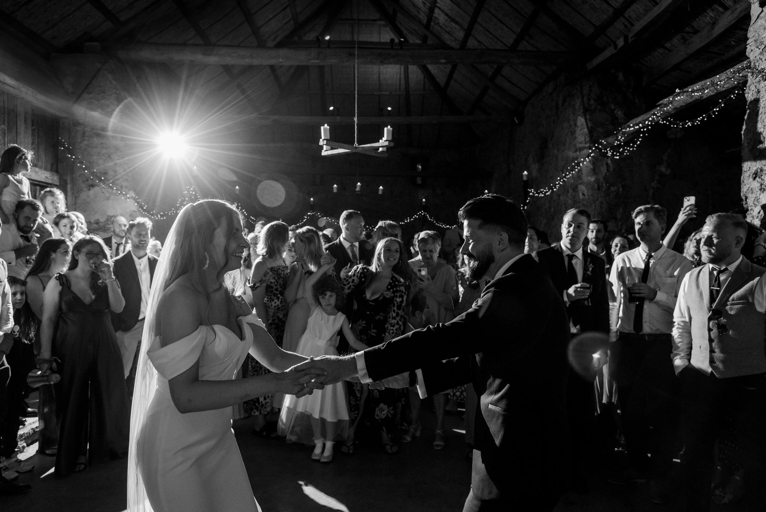 Black and white photo of a bride and groom dancing, surrounded by guests, indoors at a wedding reception with fairy lights.