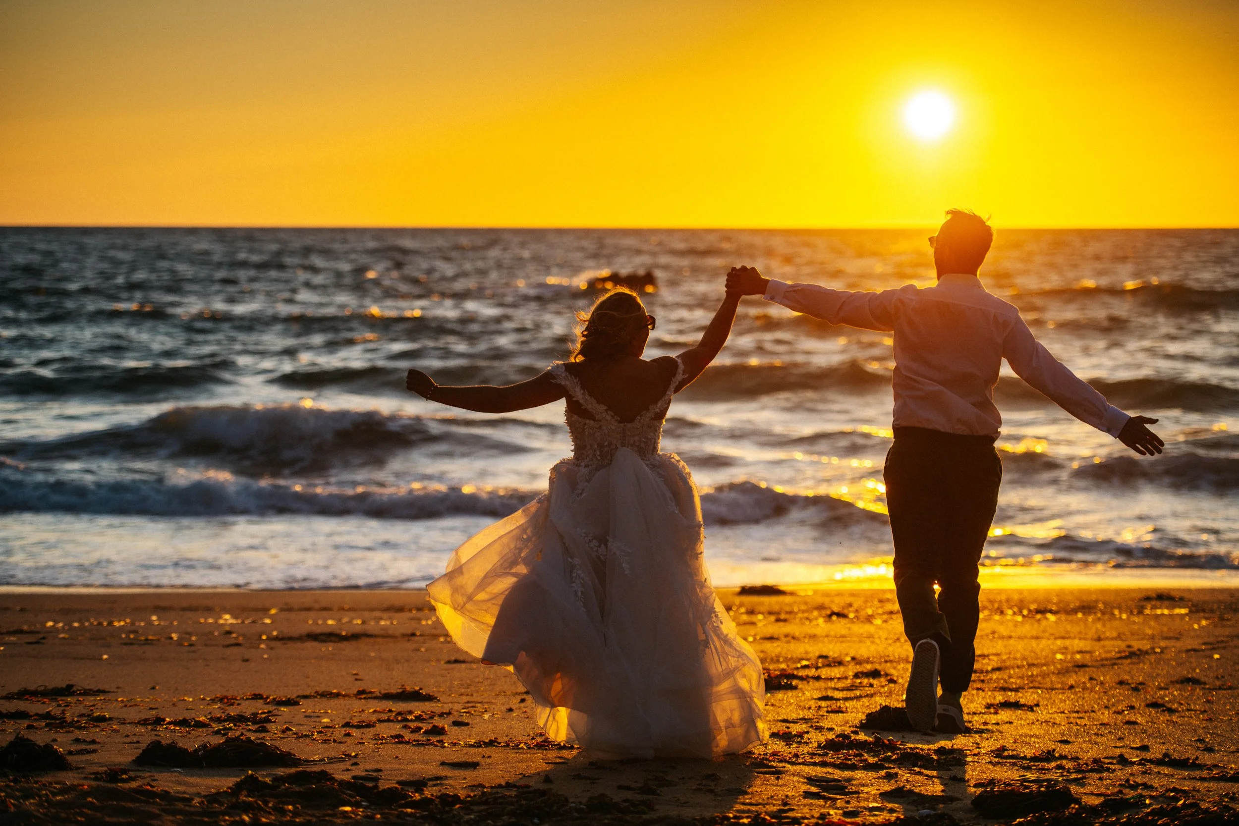 Couple in wedding attire holding hands on a beach at sunset