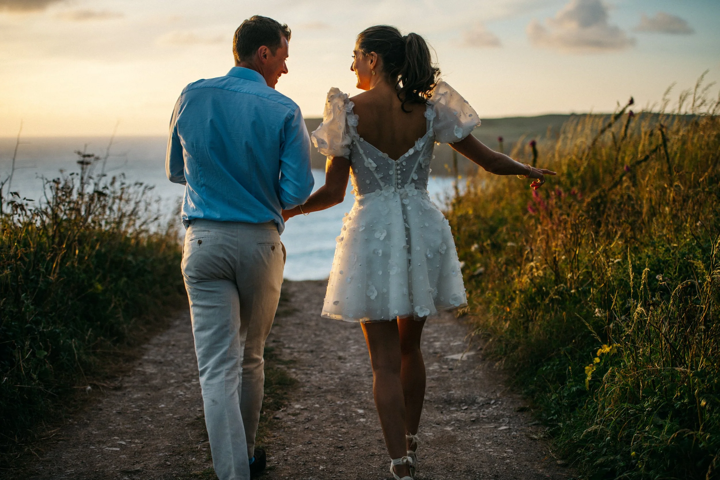 Couple walking on a path near the sea at sunset, woman in a white dress and man in light blue shirt and beige pants.
