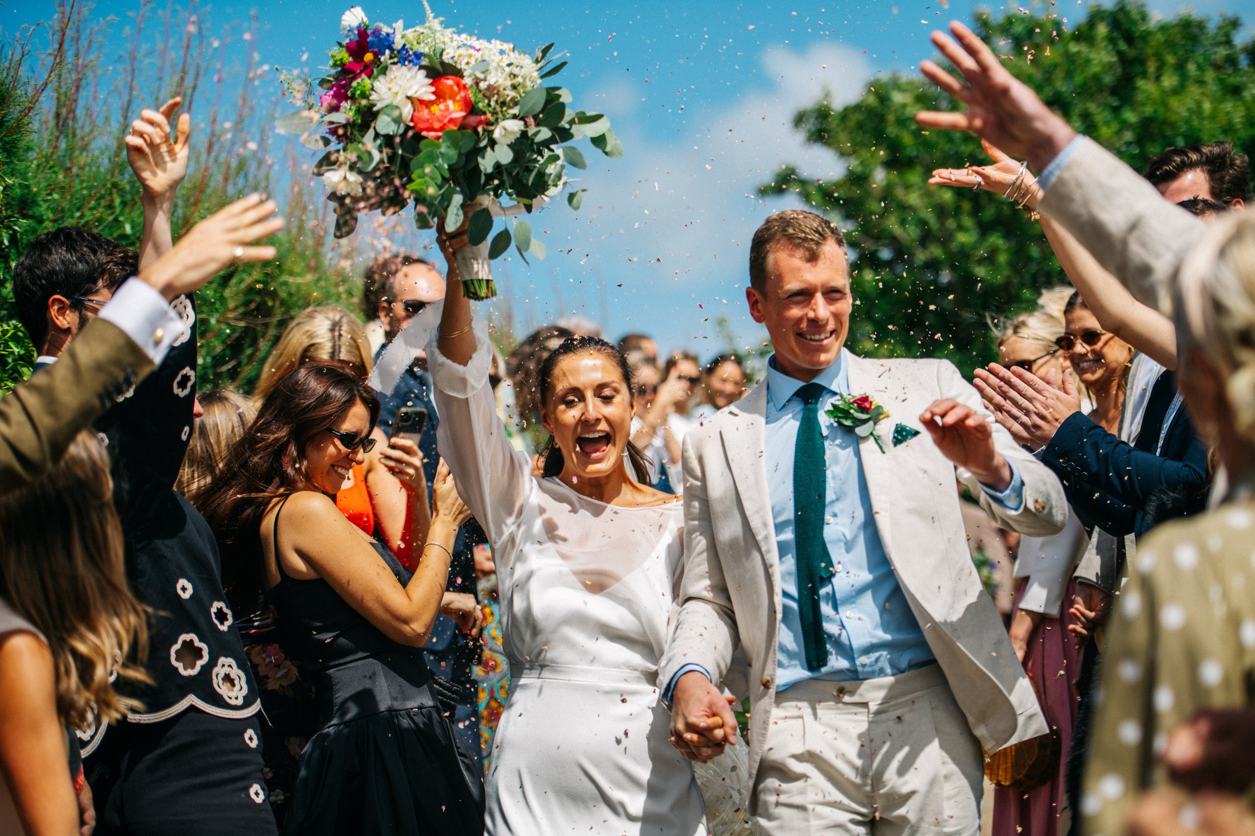 Bride and groom outside St Enodoc Church in Cornwall celebrating at outdoor wedding with bouquet and confetti.