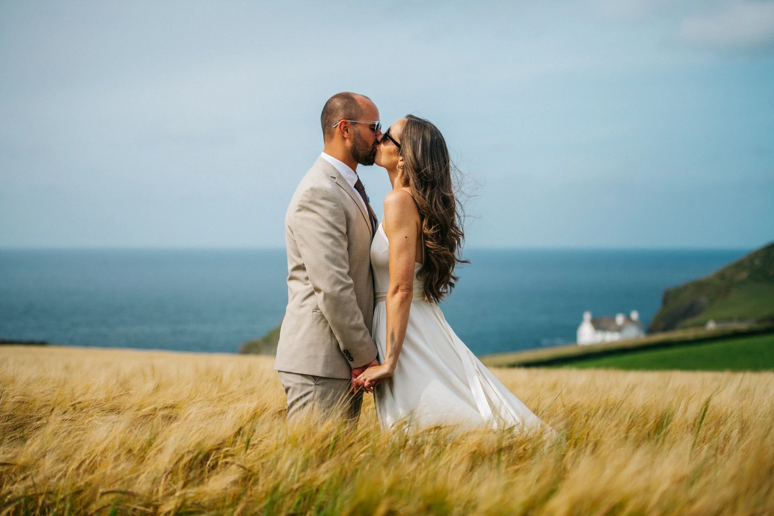 Couple in wedding attire kissing in a field near the ocean with distant hills and a house visible.