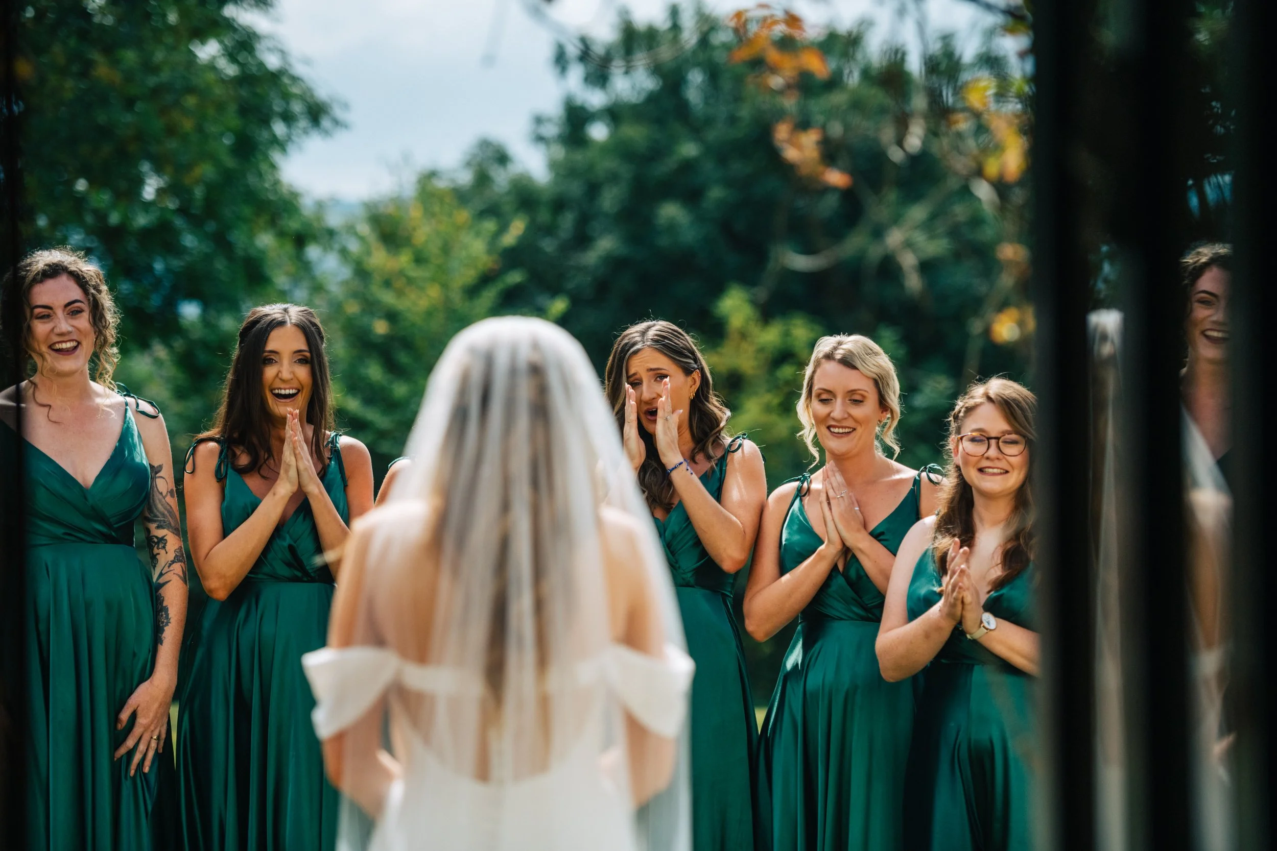 Bride with veil facing excited bridesmaids in green dresses outdoors.