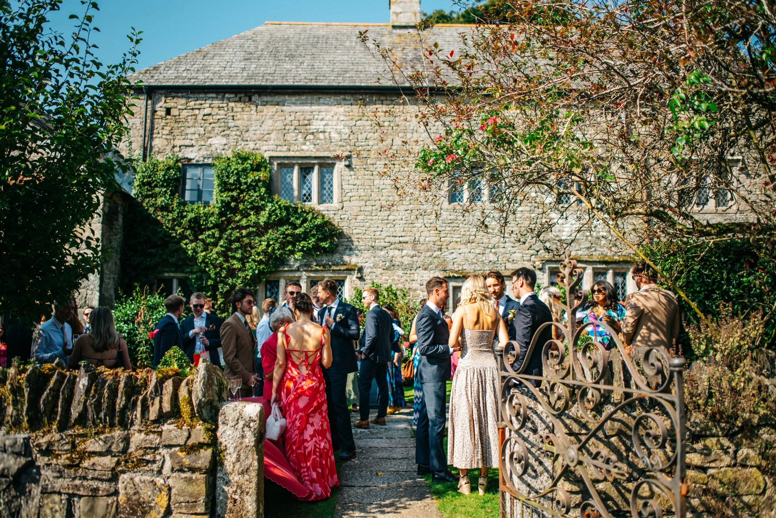Outdoor garden wedding reception at The Great Barn in Devon with people in formal attire near rustic stone building.