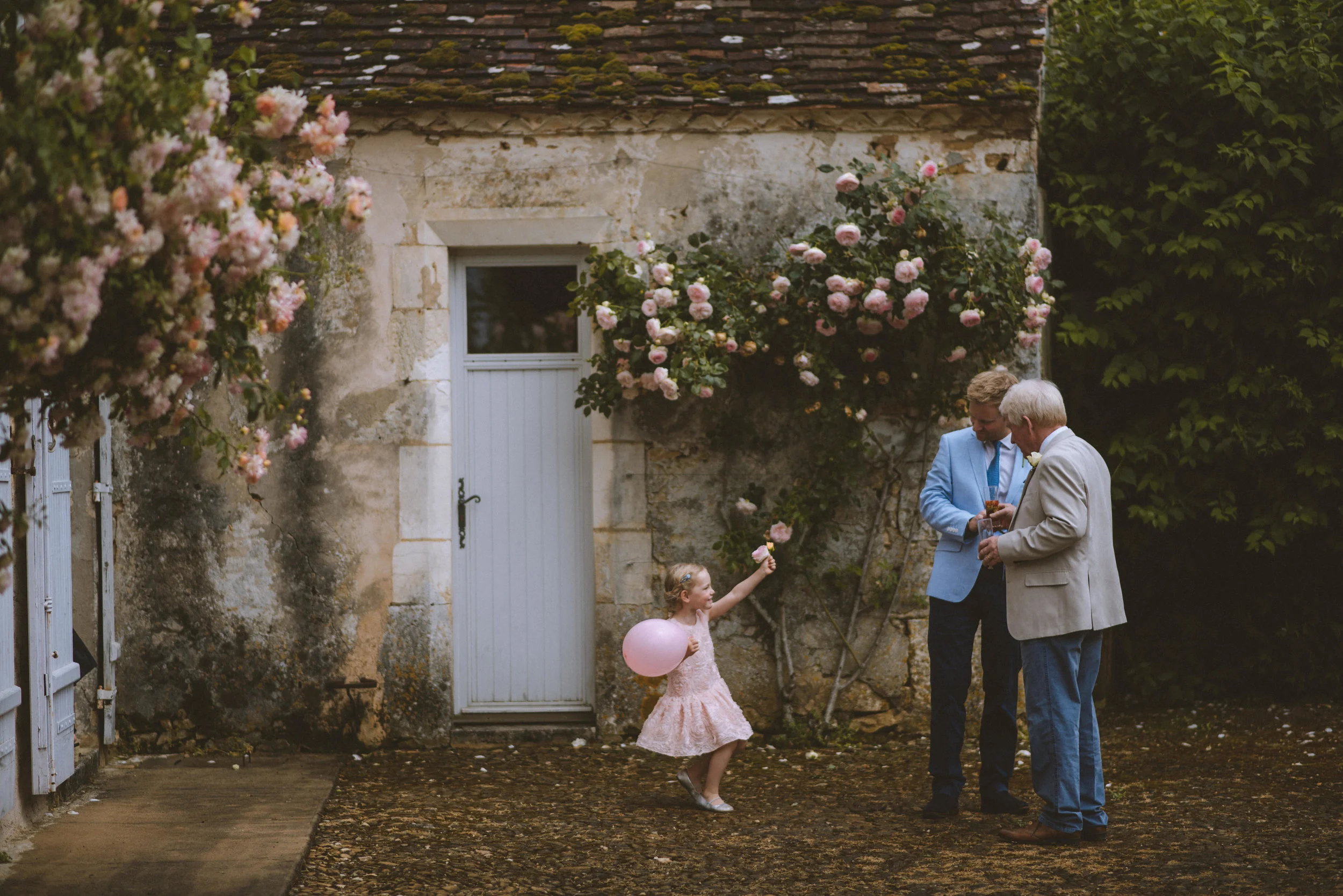 A young girl in a pink dress holding a pink balloon, reaching out to pink roses on a vine, while two older men in suits, one blonde and one with gray hair, stand nearby looking at a phone, in front of an old stone building with roses and greenery.
