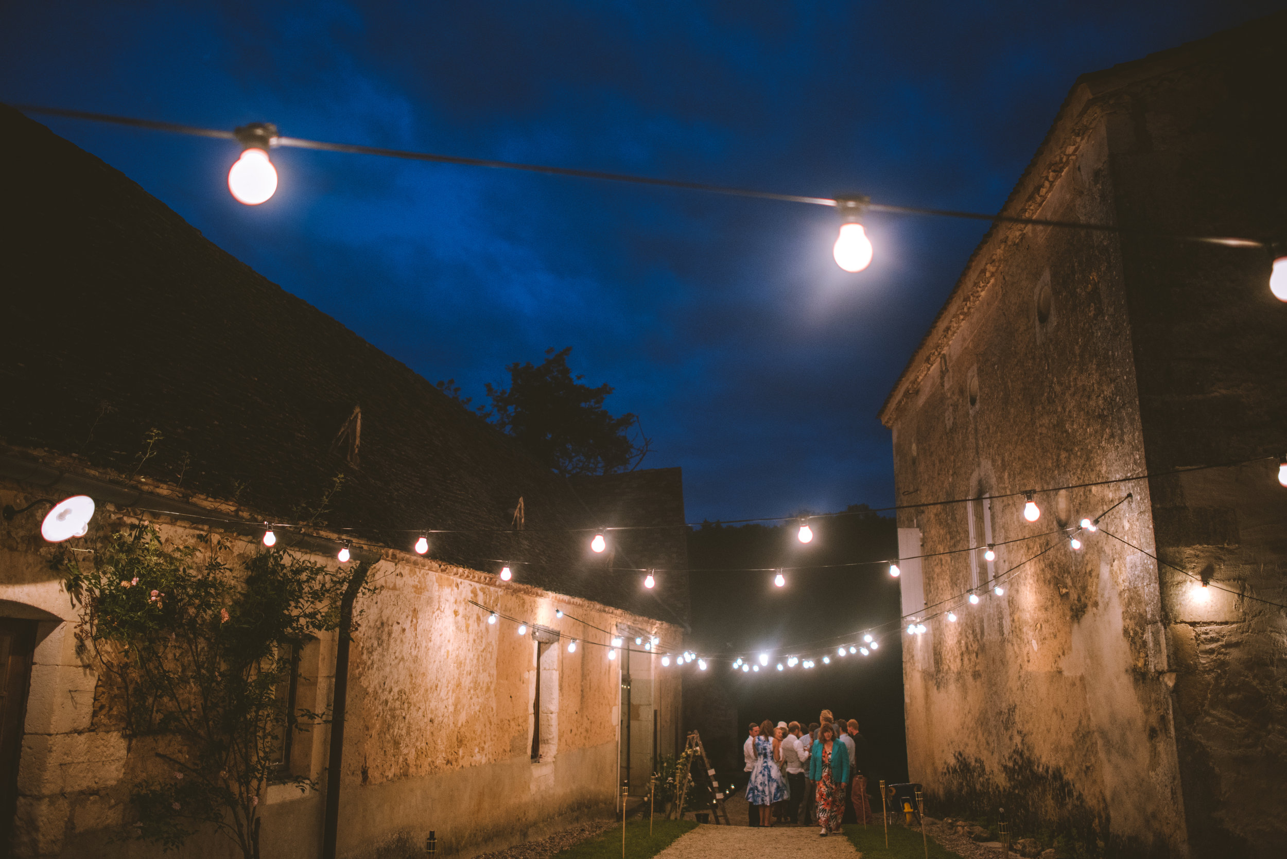 Group of people gathered together outdoors at night, under string lights, near an old stone building.