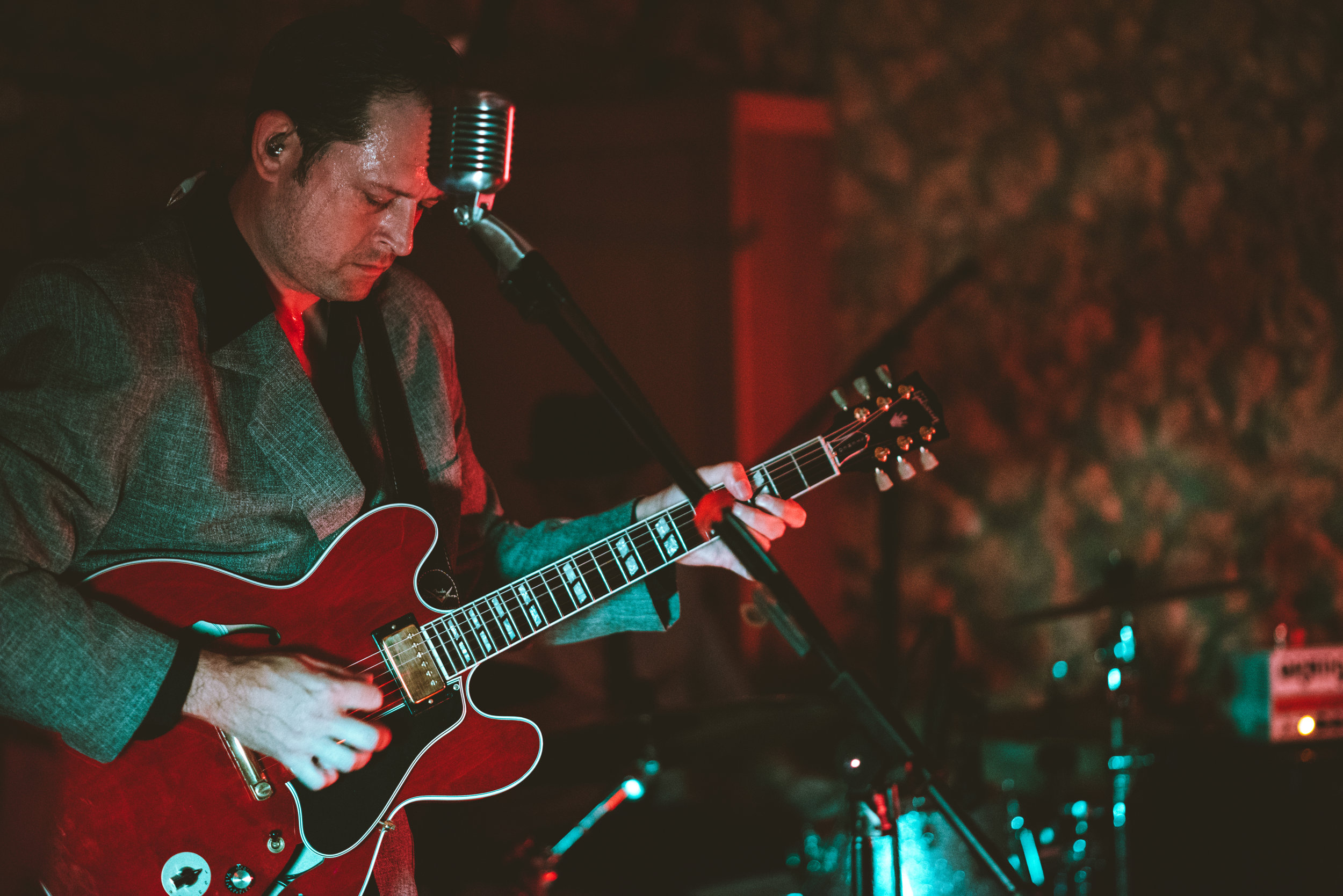 A man in a gray blazer playing a red electric guitar and singing into a vintage microphone during a live music performance.