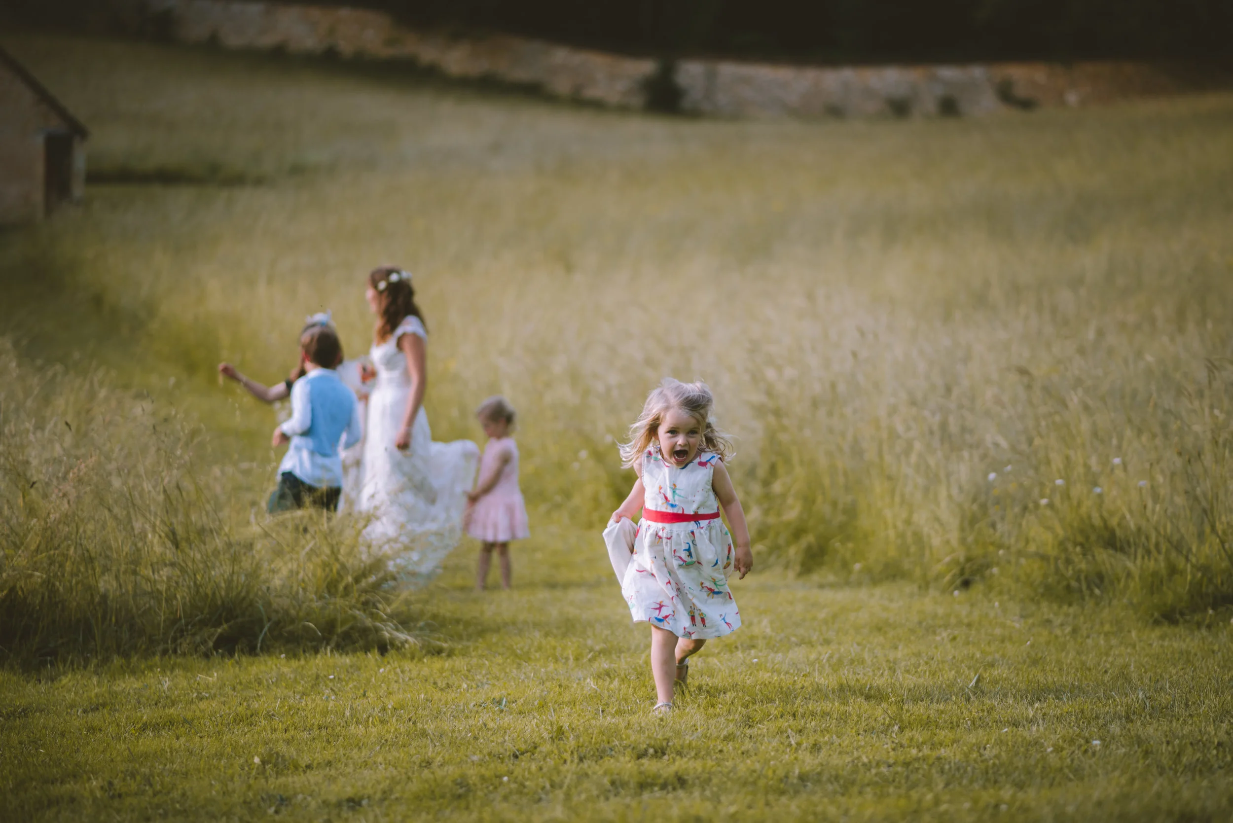 A young girl running and smiling in a grassy field with a group of people, including children and a woman, in the background.