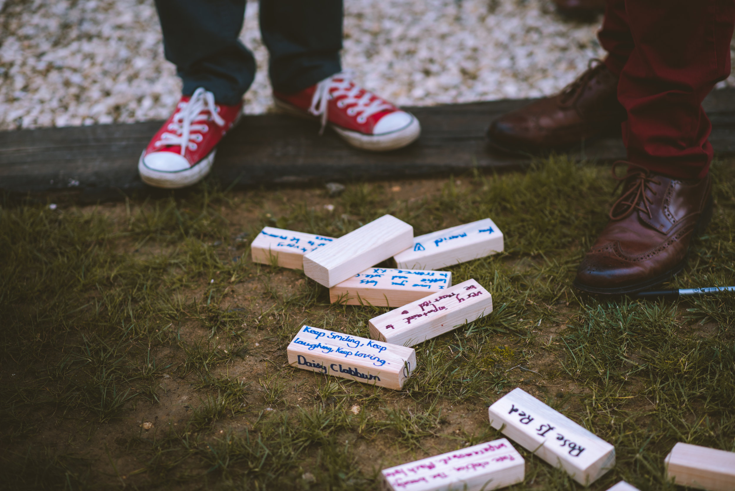 Colorfully decorated wooden blocks on the ground with messages, surrounded by two pairs of legs and shoes of different styles and colors, with grass and gravel in the background.