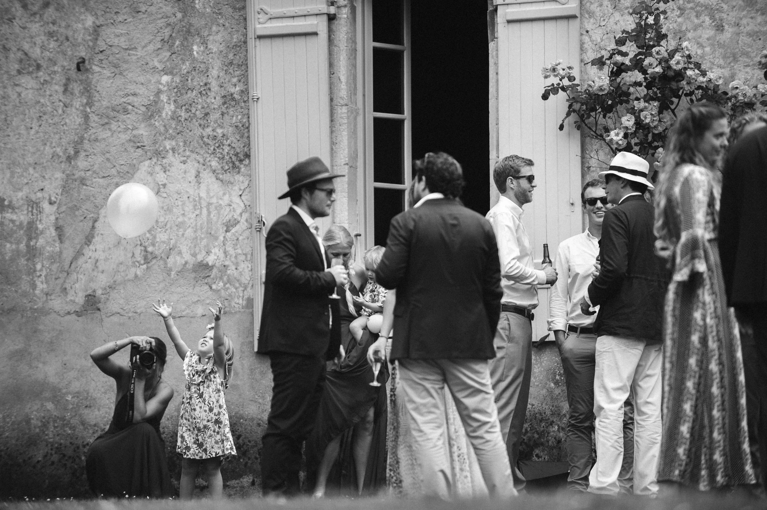 Group of people at outdoor gathering, some holding drinks, a woman taking a photo of a young girl reaching for a balloon, with an old stone wall and open window in the background.