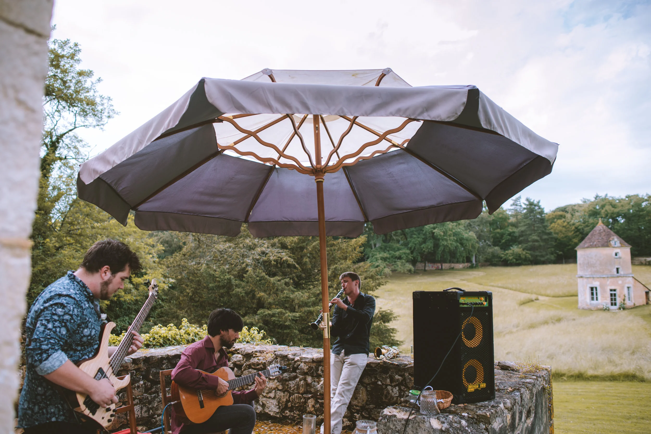 three musicians playing instruments outdoors beneath a large umbrella, with a stone wall and a scenic landscape of grassy fields, trees, and a small building in the background.