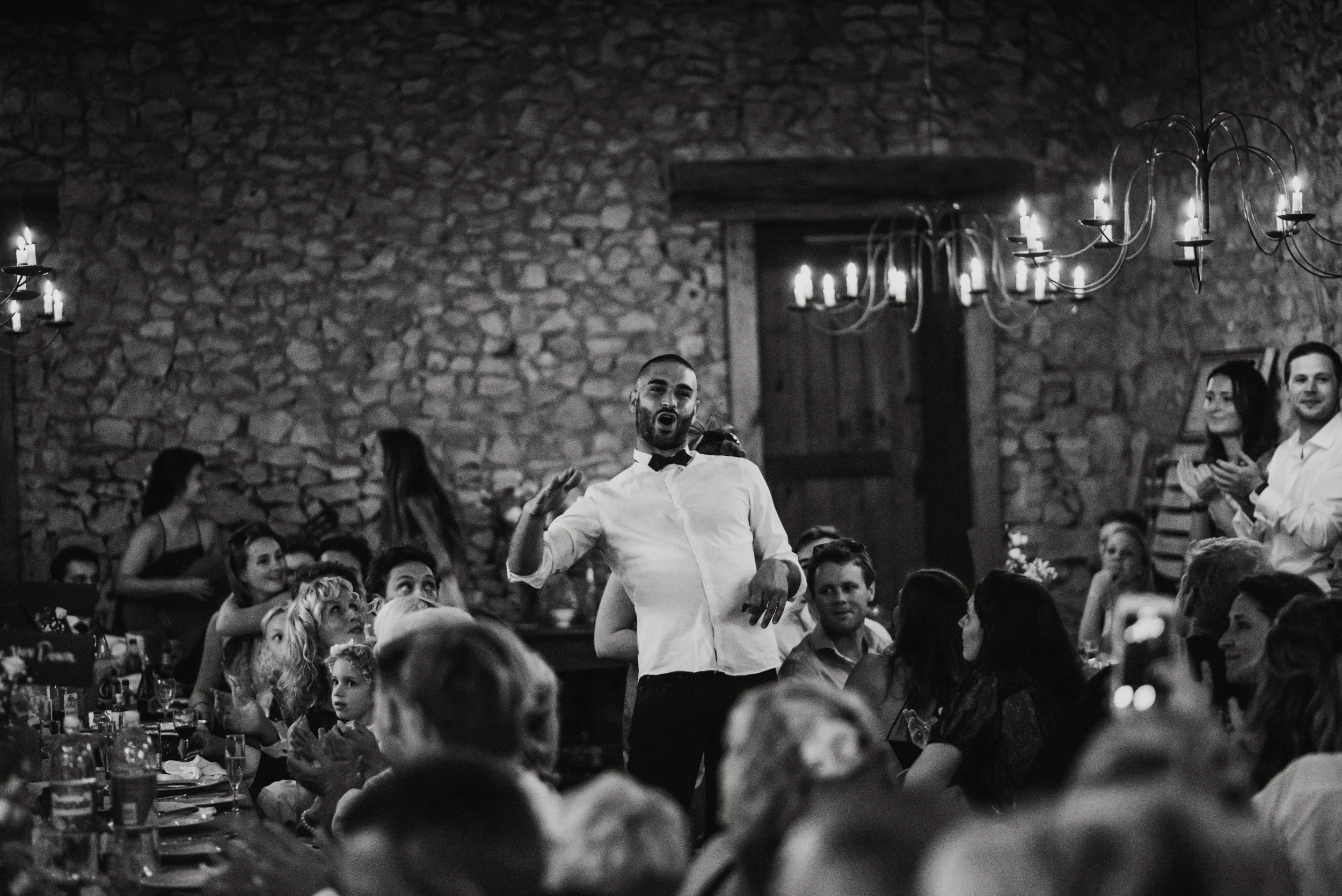 A man with a beard in a white shirt and bow tie stands in front of a crowd at a celebration, possibly a wedding reception, with stone walls and chandeliers in the background. The image is in black and white.