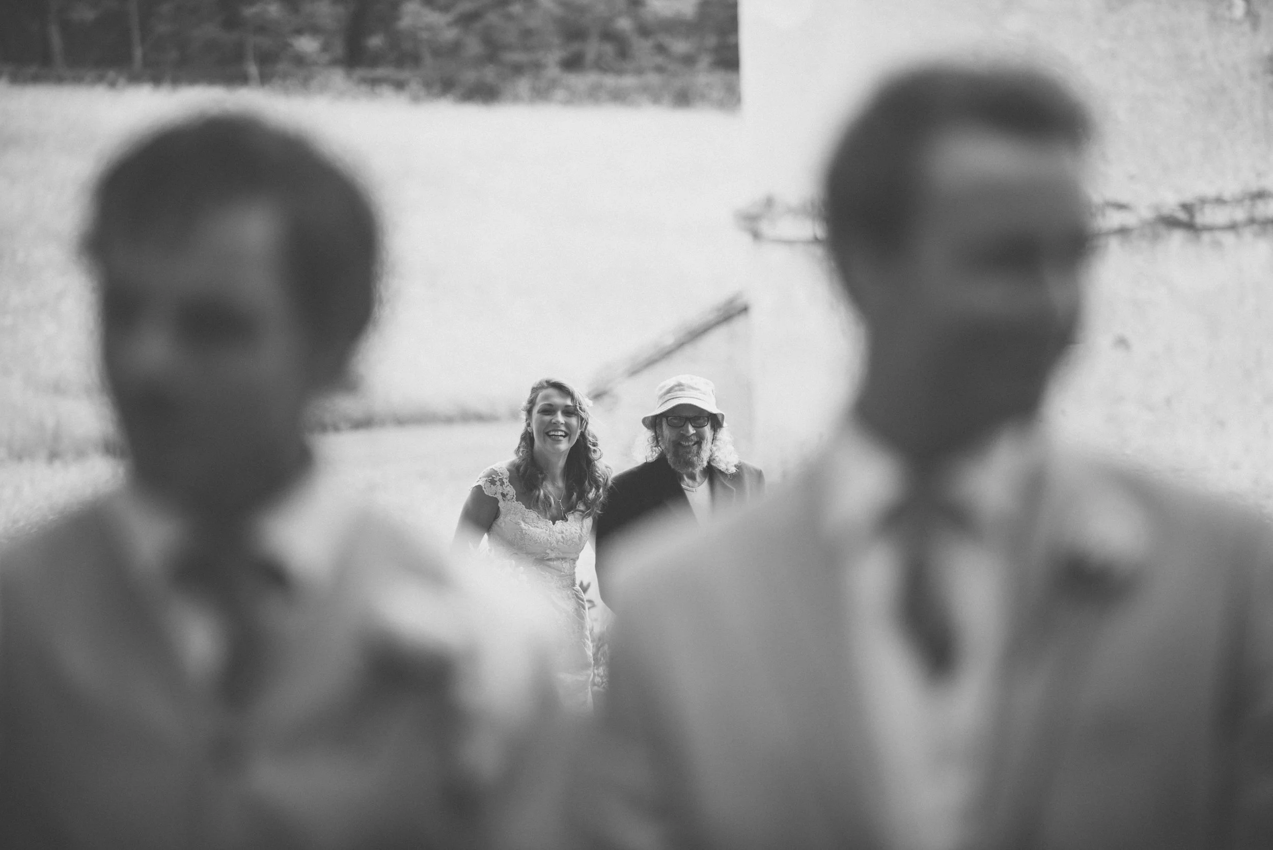 Black and white photo of a wedding scene with the bride and a man behind the blurred figures of two other men in suits and ties in the foreground.