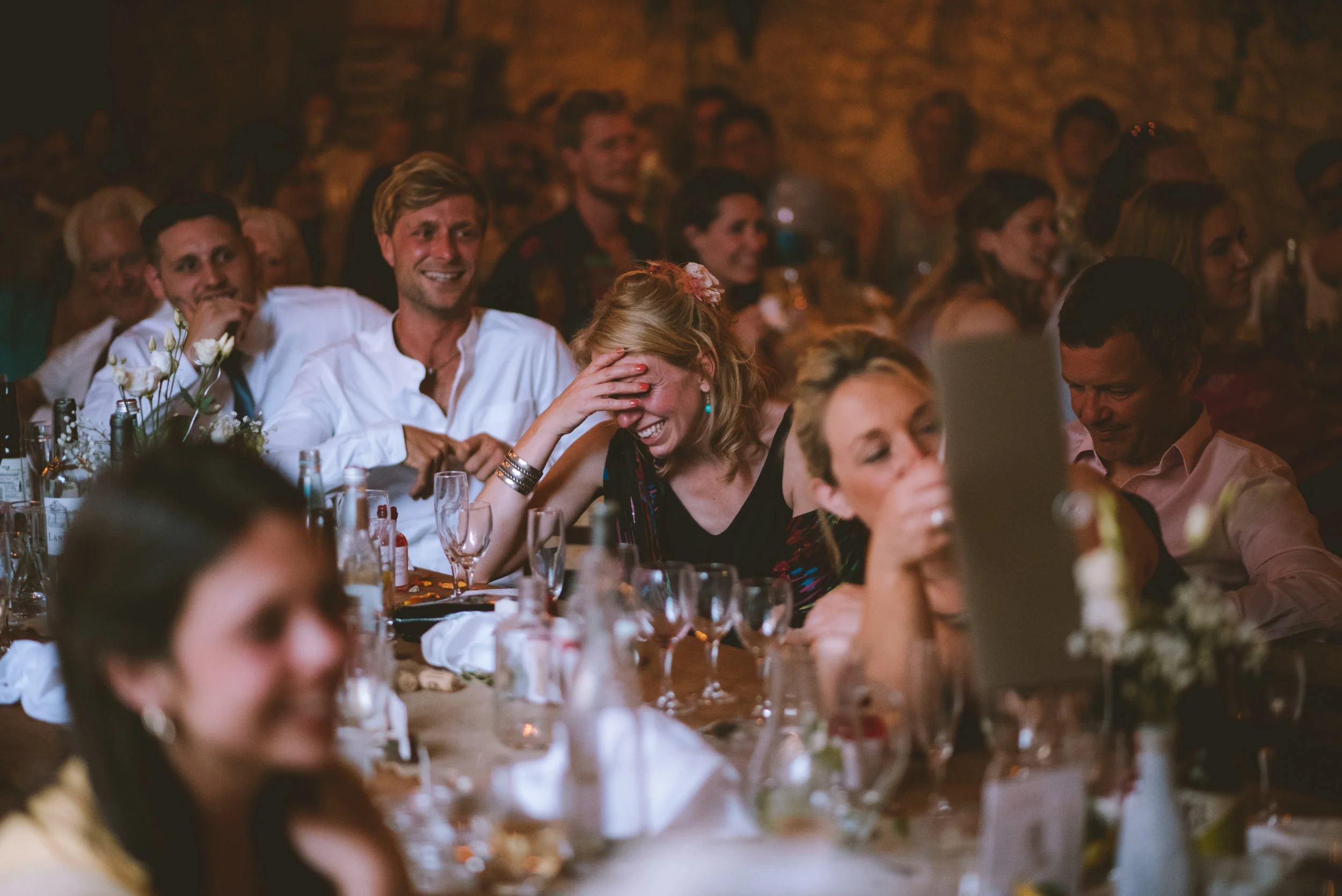 People sitting at a table during a celebration, smiling and laughing, with a woman covering her face in joy.