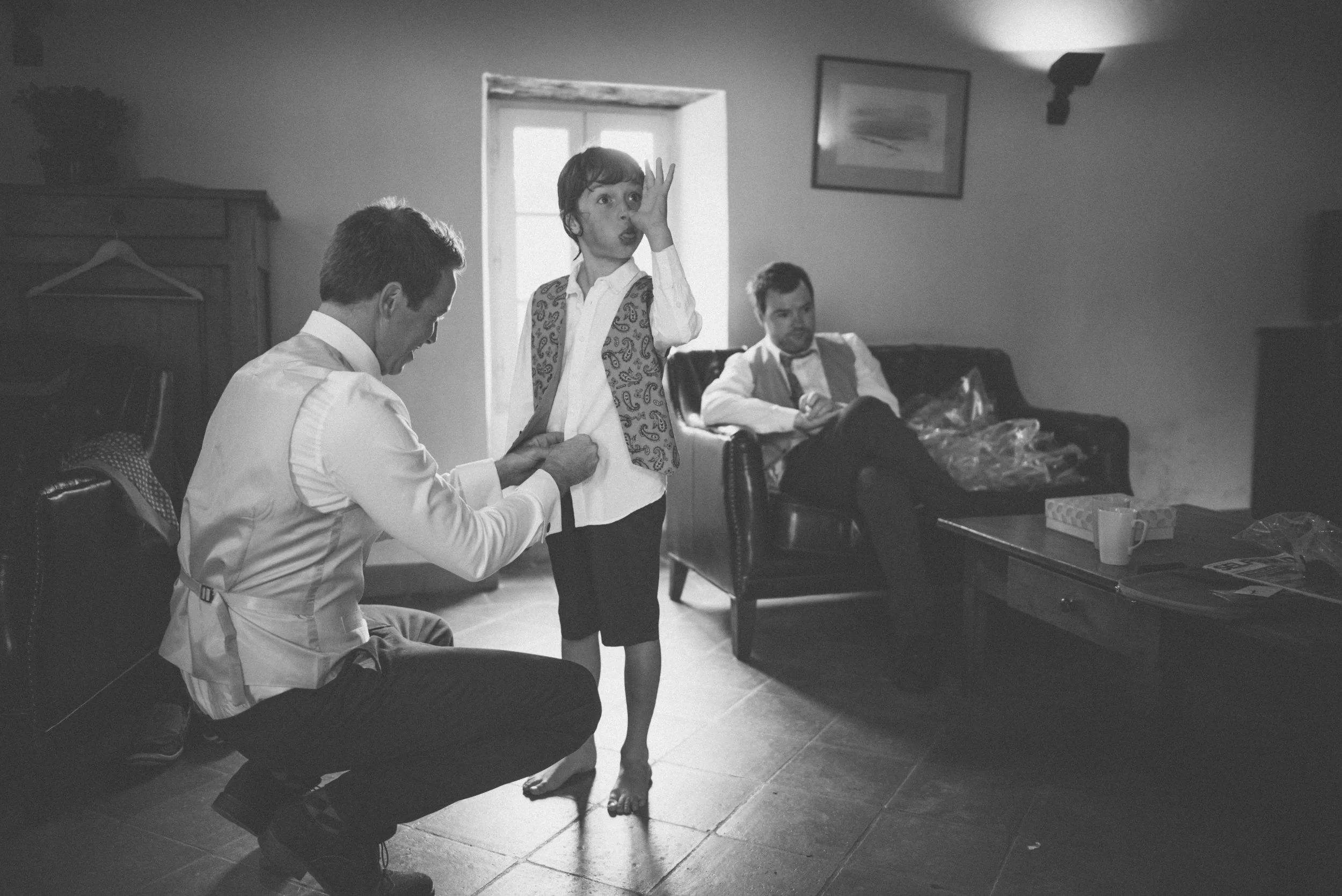 A black and white photo of a young boy holding his nose as an older man, possibly a doctor, adjusts the boy's shirt collar. An adult man, possibly a father, sits on a sofa in the background.