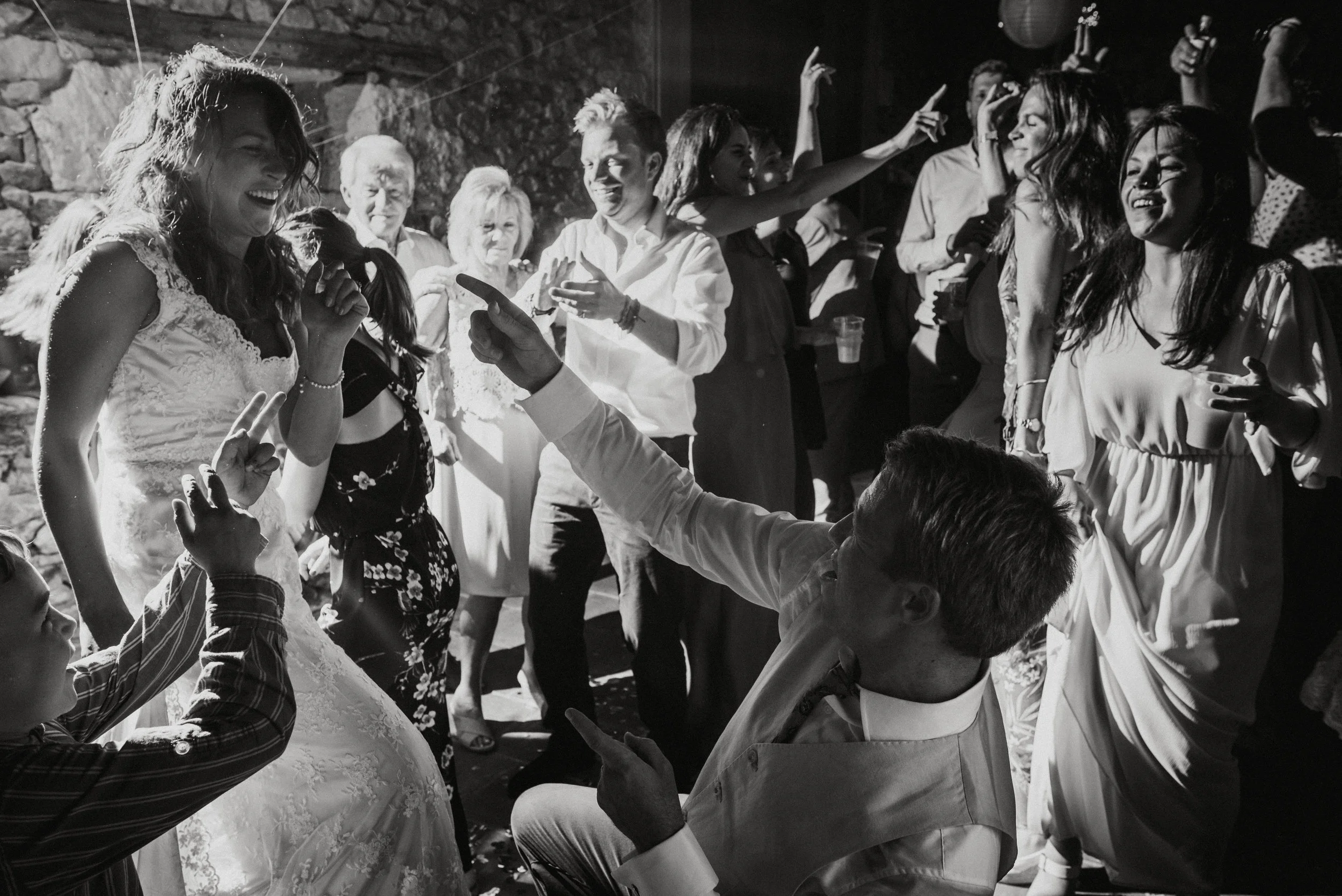 Black-and-white photo of a lively celebration with a woman in a wedding dress, surrounded by friends and family dancing and smiling.